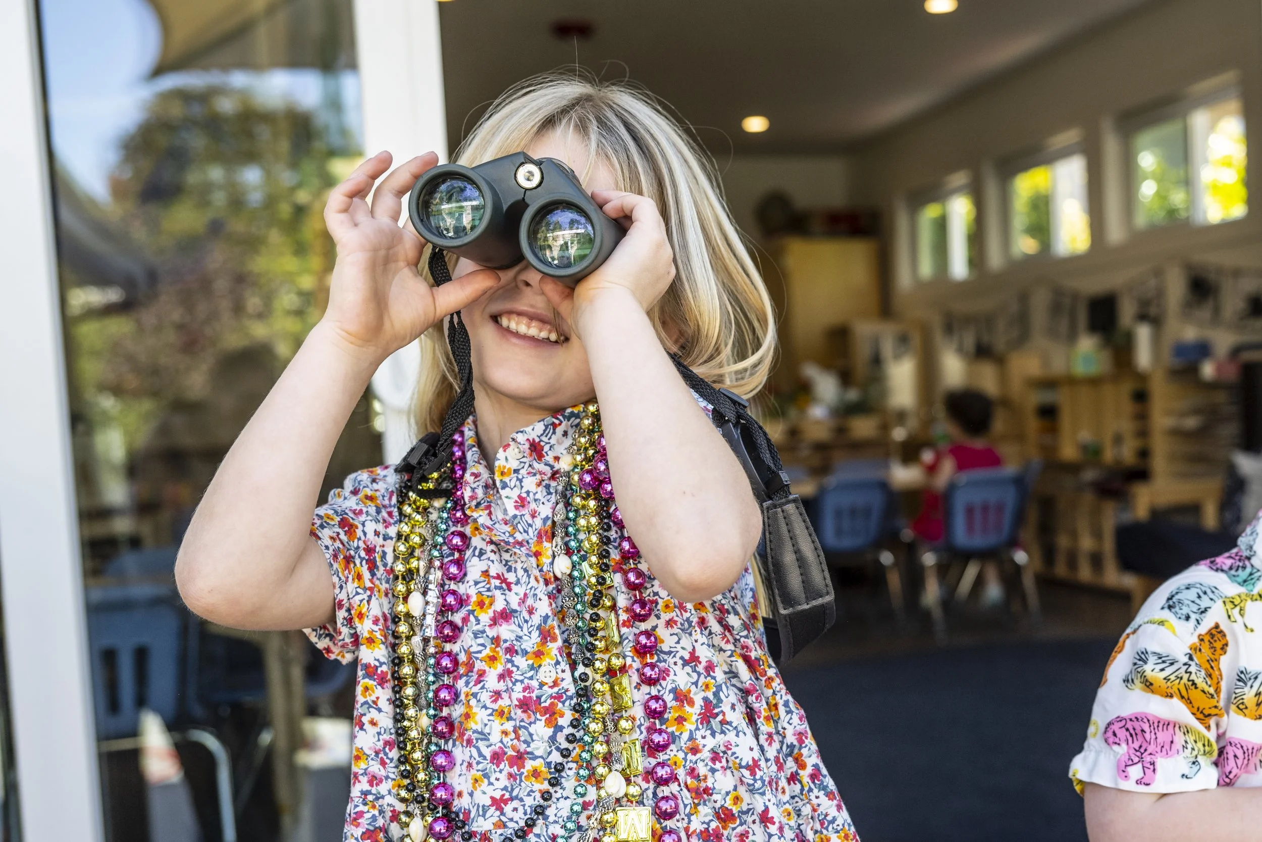 A young girl with blonde hair wearing a floral dress and colorful beads around her neck is looking through binoculars indoors near a large window, with another child visible in the background.