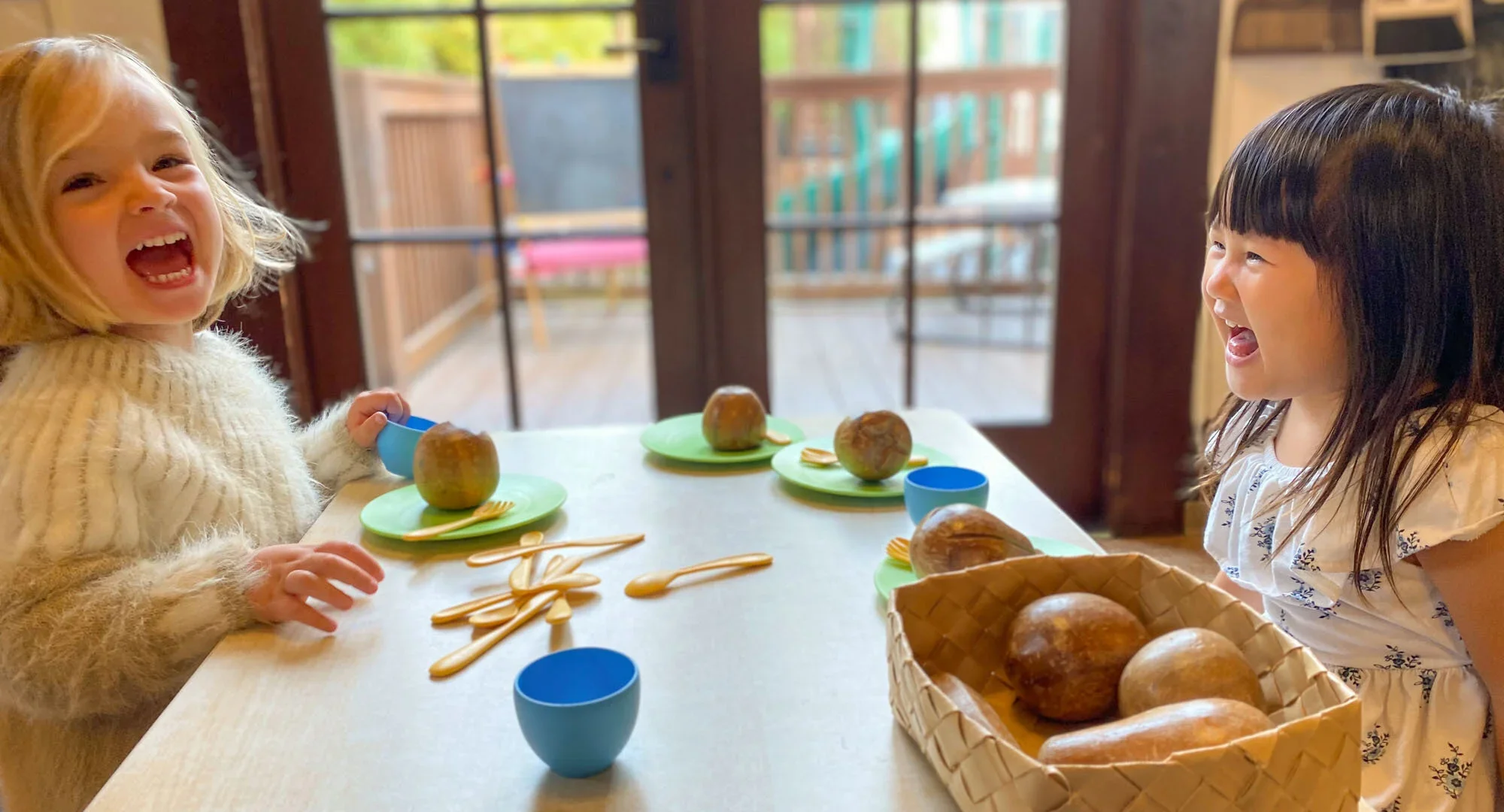 Two young girls sitting at a table, laughing and playing with toy food and utensils, with a basket of real bread on the table, in a room with glass doors leading to an outdoor deck.