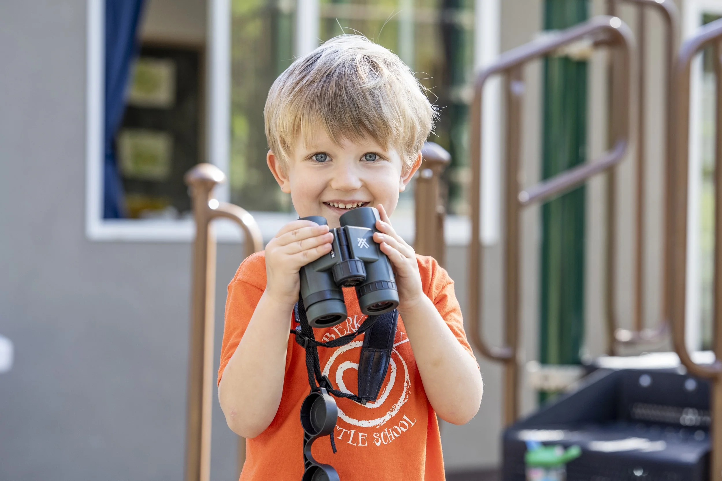 A young boy with blonde hair and blue eyes holding binoculars and smiling.