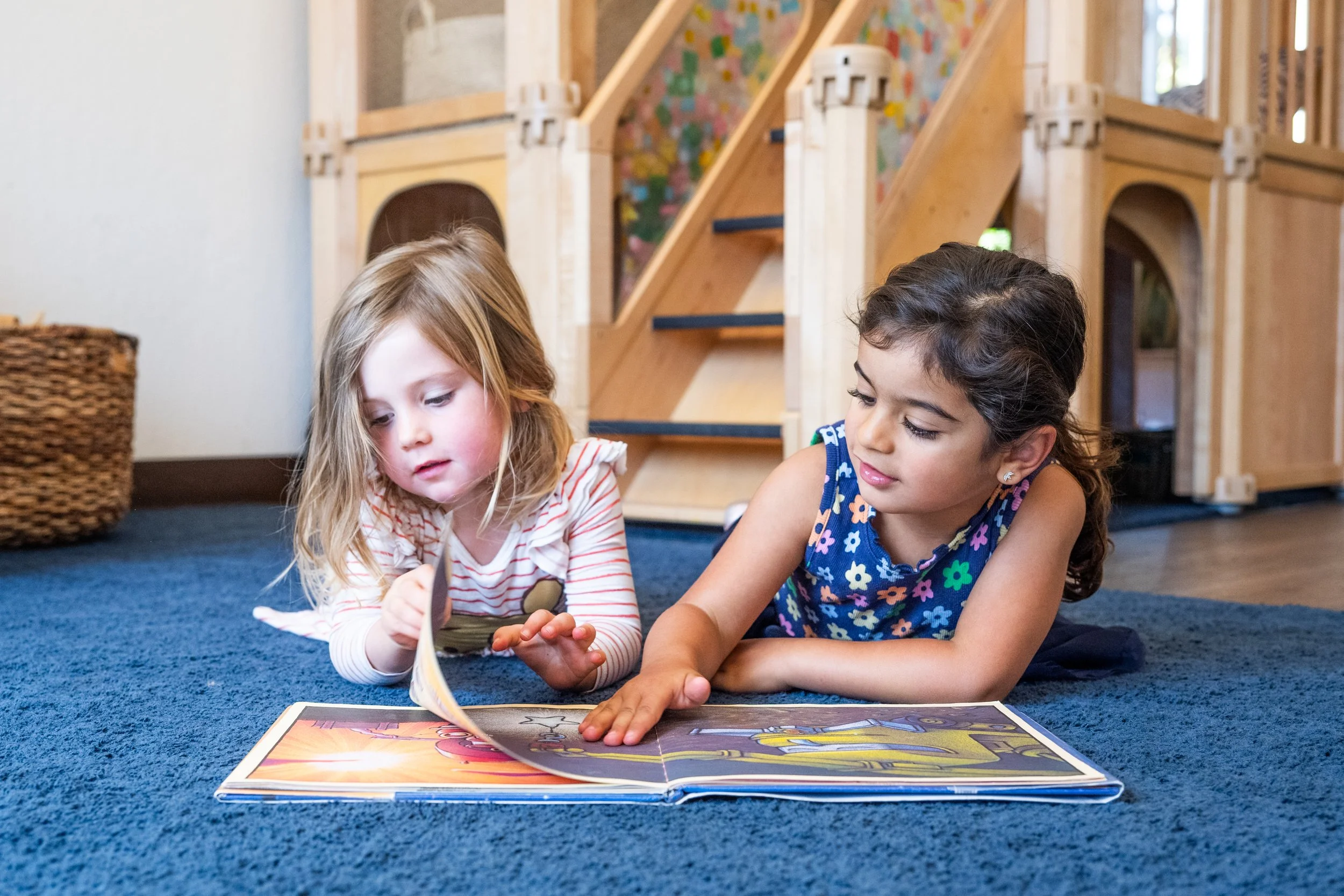 Two young girls lying on a blue carpet, looking at a large illustrated book, with a wooden playhouse structure in the background.