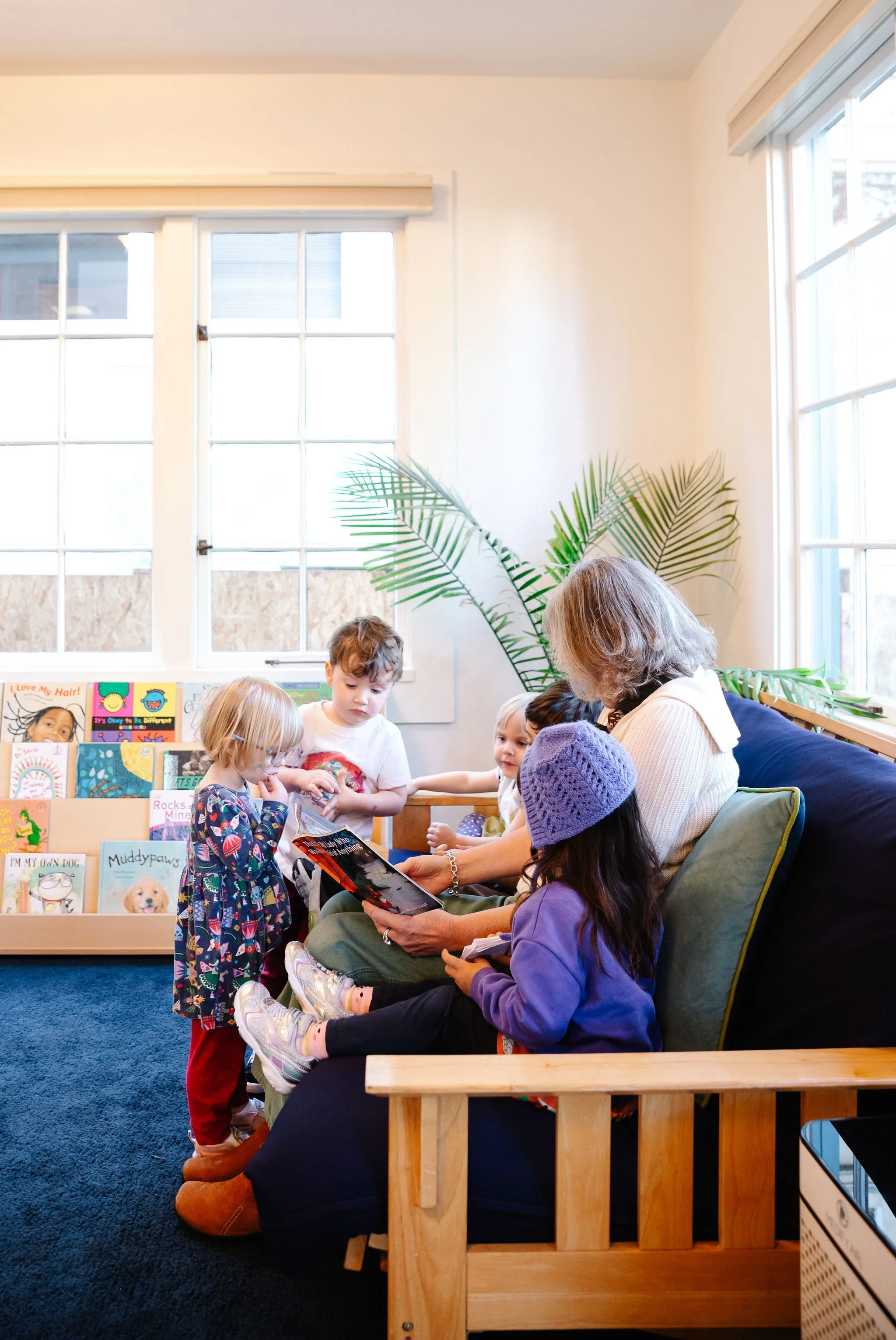 A woman reading a book to five children in a cozy, brightly lit room with large windows, houseplants, and a bookshelf with children's books.