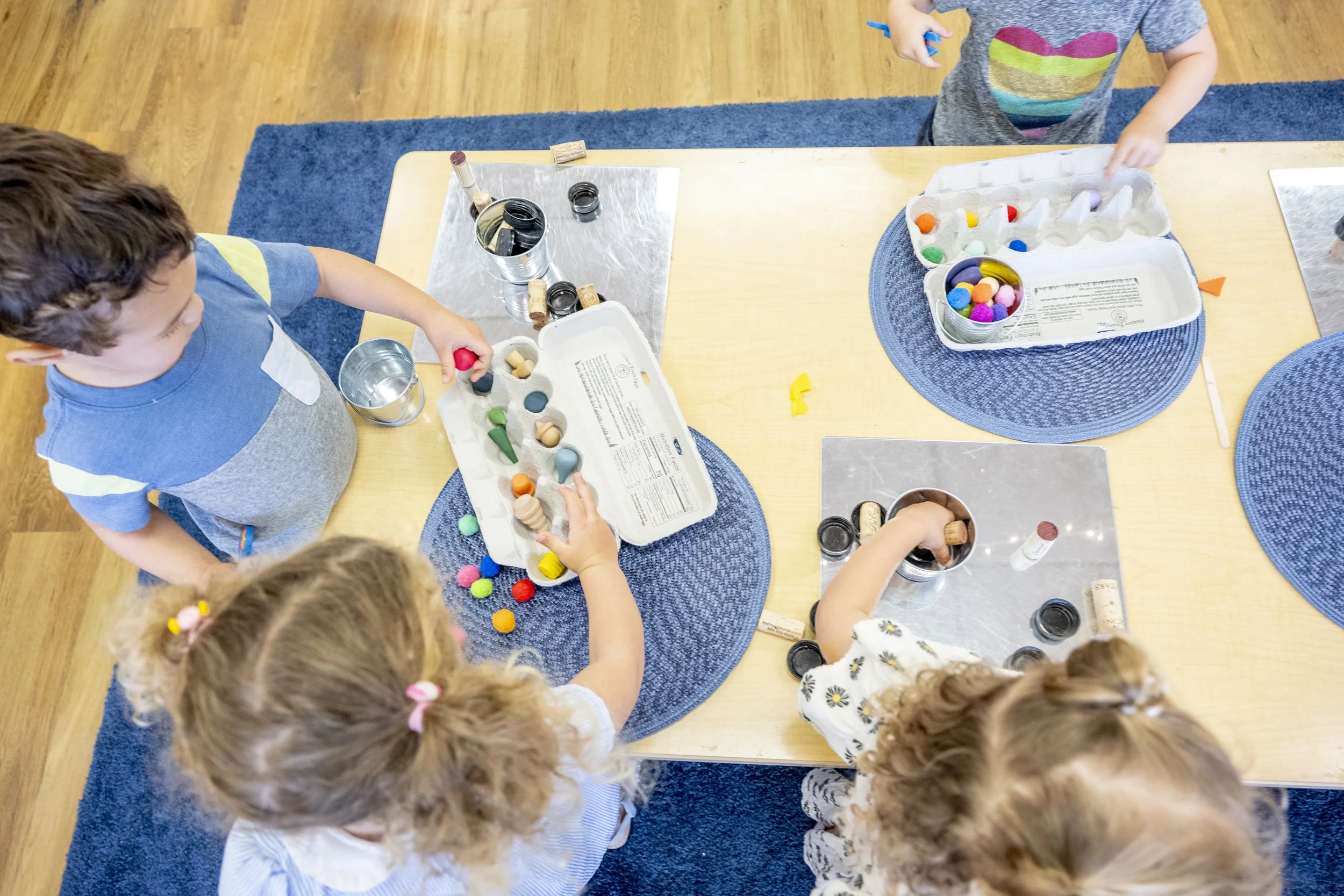 Children engaging in a craft activity at a table with colorful pom-poms, corks, and small containers.
