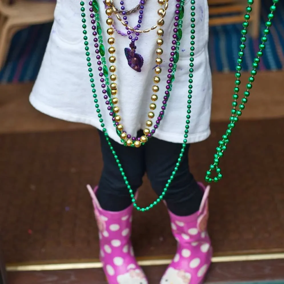 Child wearing colorful bead necklaces, pink rain boots with white polka dots, black pants, and a white shirt, standing on a wooden surface.