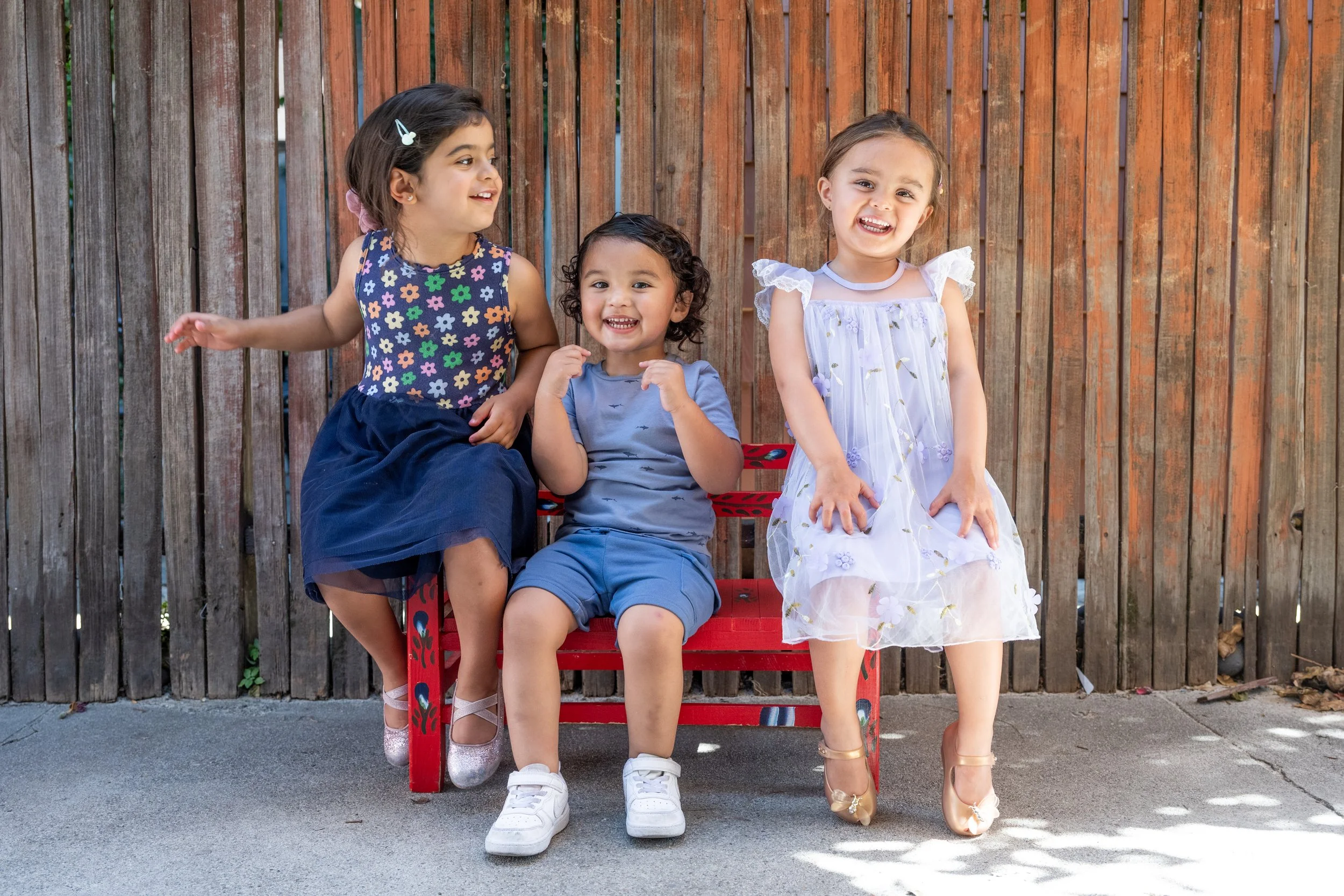Three young girls sitting on a red bench against a wooden fence, smiling and enjoying each other's company.