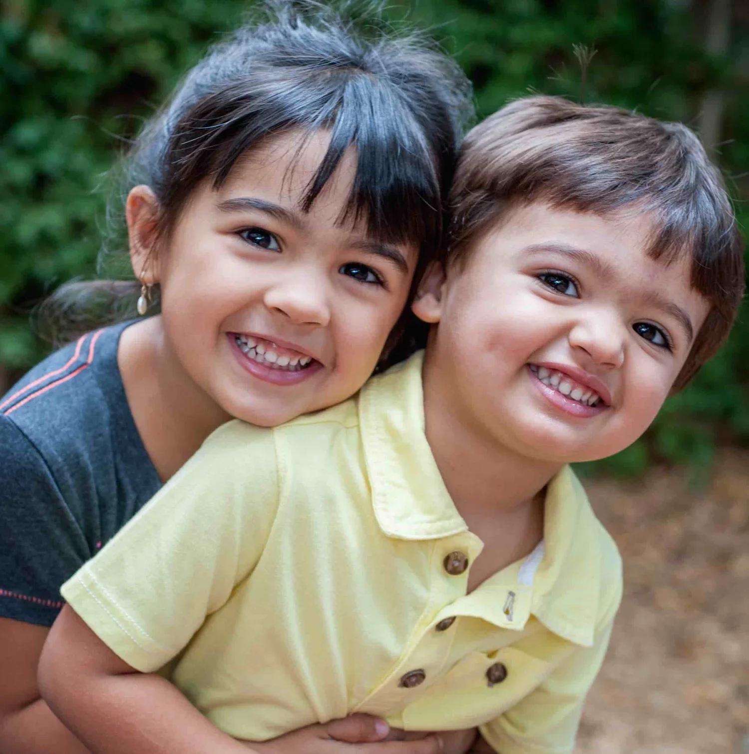 Two smiling children, a girl and a boy, close together outdoors with greenery in the background.