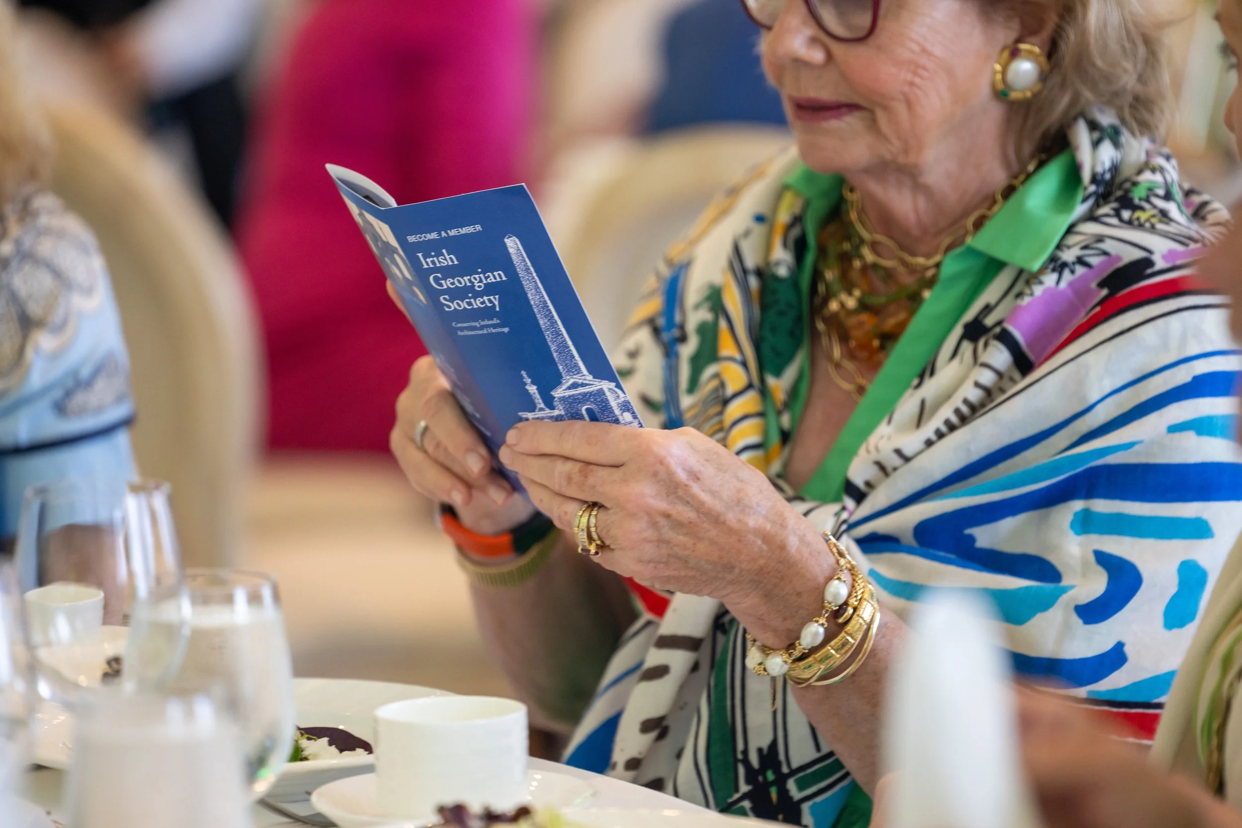 An elderly woman with blonde hair, wearing glasses, colorful jewelry, and a multicolored striped jacket, holding a blue brochure for the Irish Georgian Society while seated at a table with desserts and wine glasses.