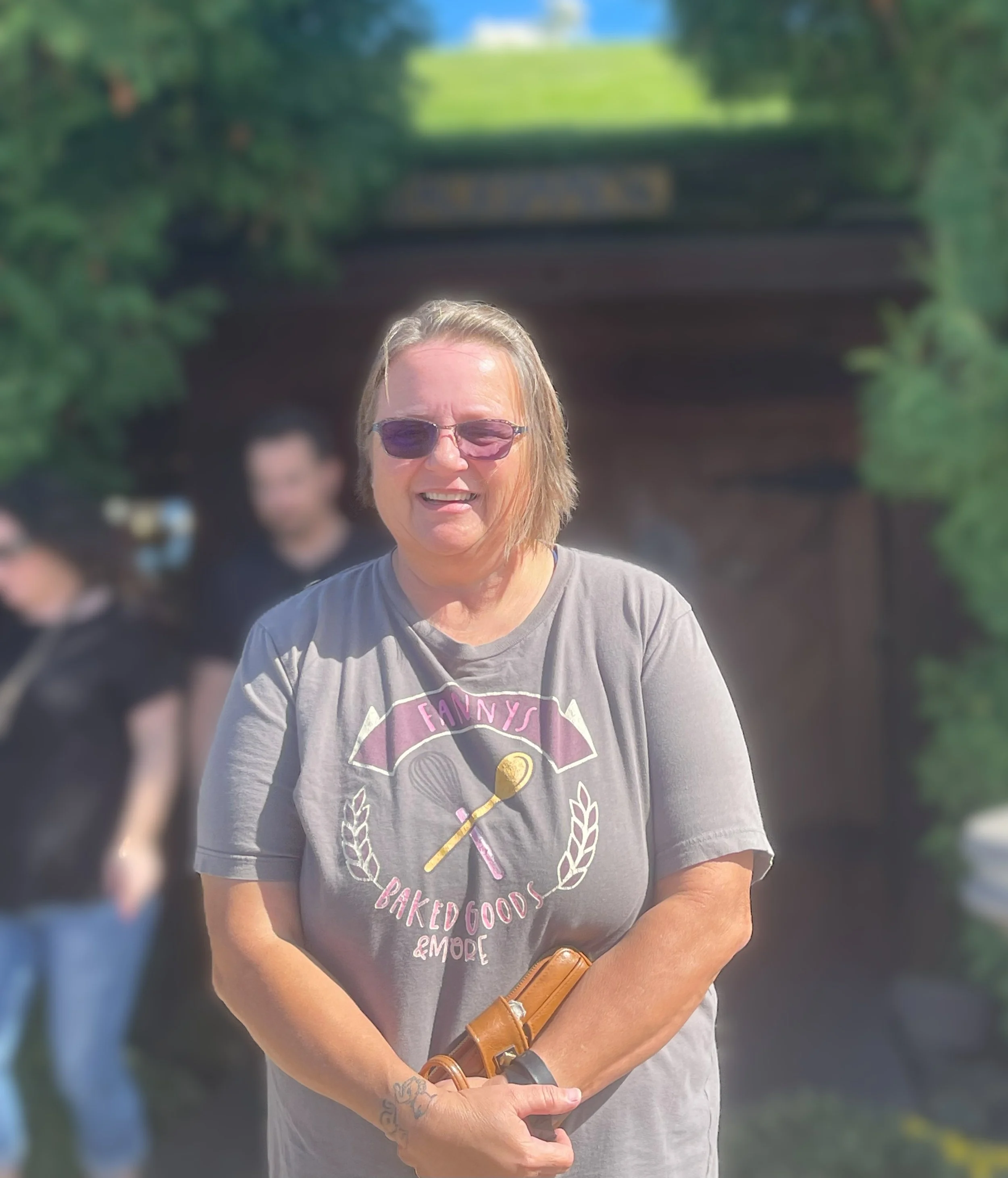 A woman with short blonde hair, wearing sunglasses and a gray T-shirt that says Fanny's Baked Goods & More, standing outdoors with a group of people blurred in the background near a wooded area and a small bridge.