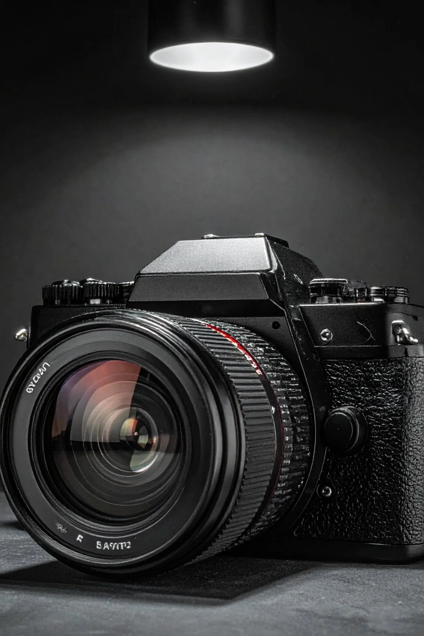 A black digital camera with a large lens on a dark surface, illuminated by a ceiling light overhead.