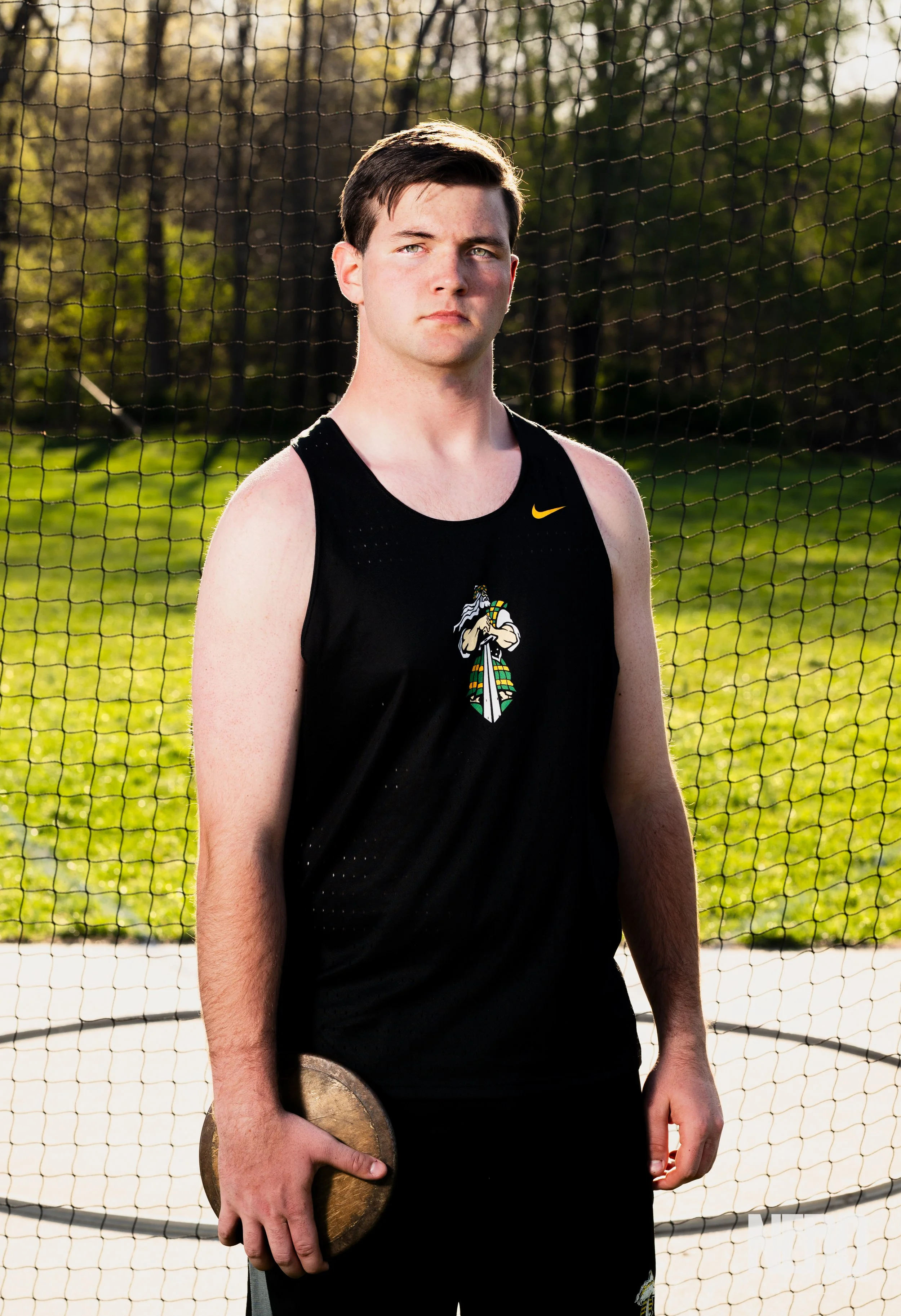 A young male athlete with short dark hair, light skin, and a serious expression, standing outdoors next to a sports net on a sunny day. He is wearing a black sleeveless sports jersey with a colorful cartoon-like Viking logo and a small yellow Nike logo. He is holding a shot put in his right hand, with a background of green grass, trees, and a partly cloudy sky.