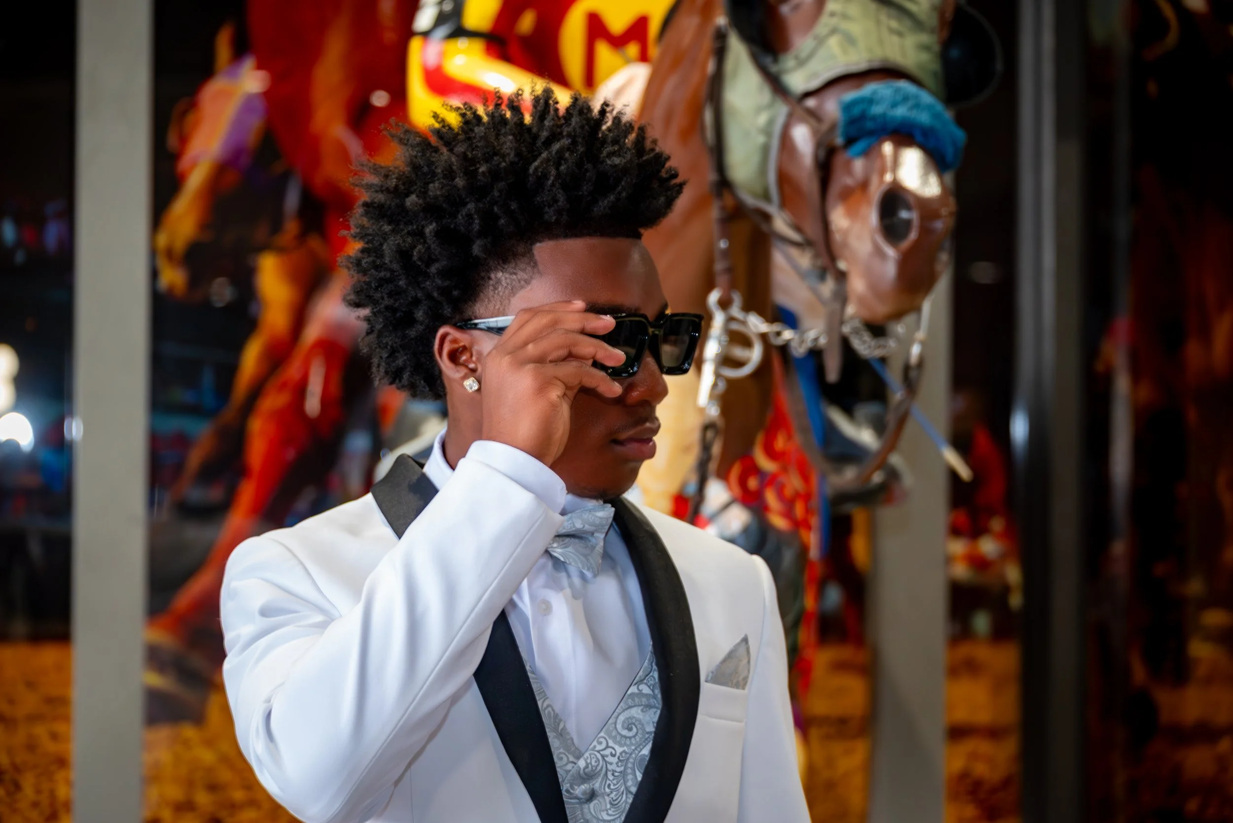 Young man in a white tuxedo with black lapels and a gray paisley vest, adjusting his sunglasses in front of carousel horses at an amusement park.