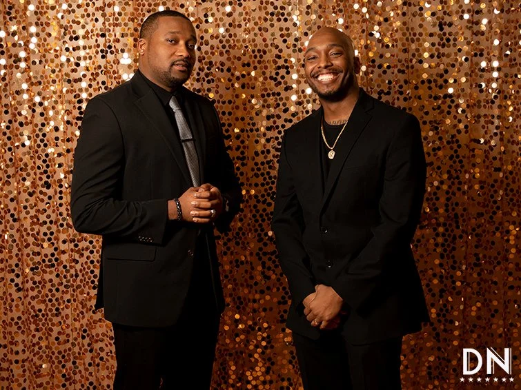 Two men in black suits standing in front of a gold sequin backdrop, smiling and posing for the photo.