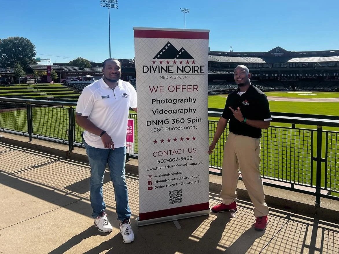 Two men standing next to a banner on a sports field. The banner advertises Divine Noire Media Group offering photography, videography, and 360 spin photobooth services, with contact information. The men are smiling and wearing casual polo shirts and pants, standing outdoors in a stadium with a baseball field in the background.