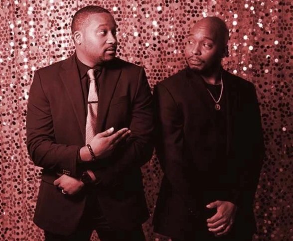Two men in black suits standing in front of a pink and gold sequin backdrop, engaged in conversation.