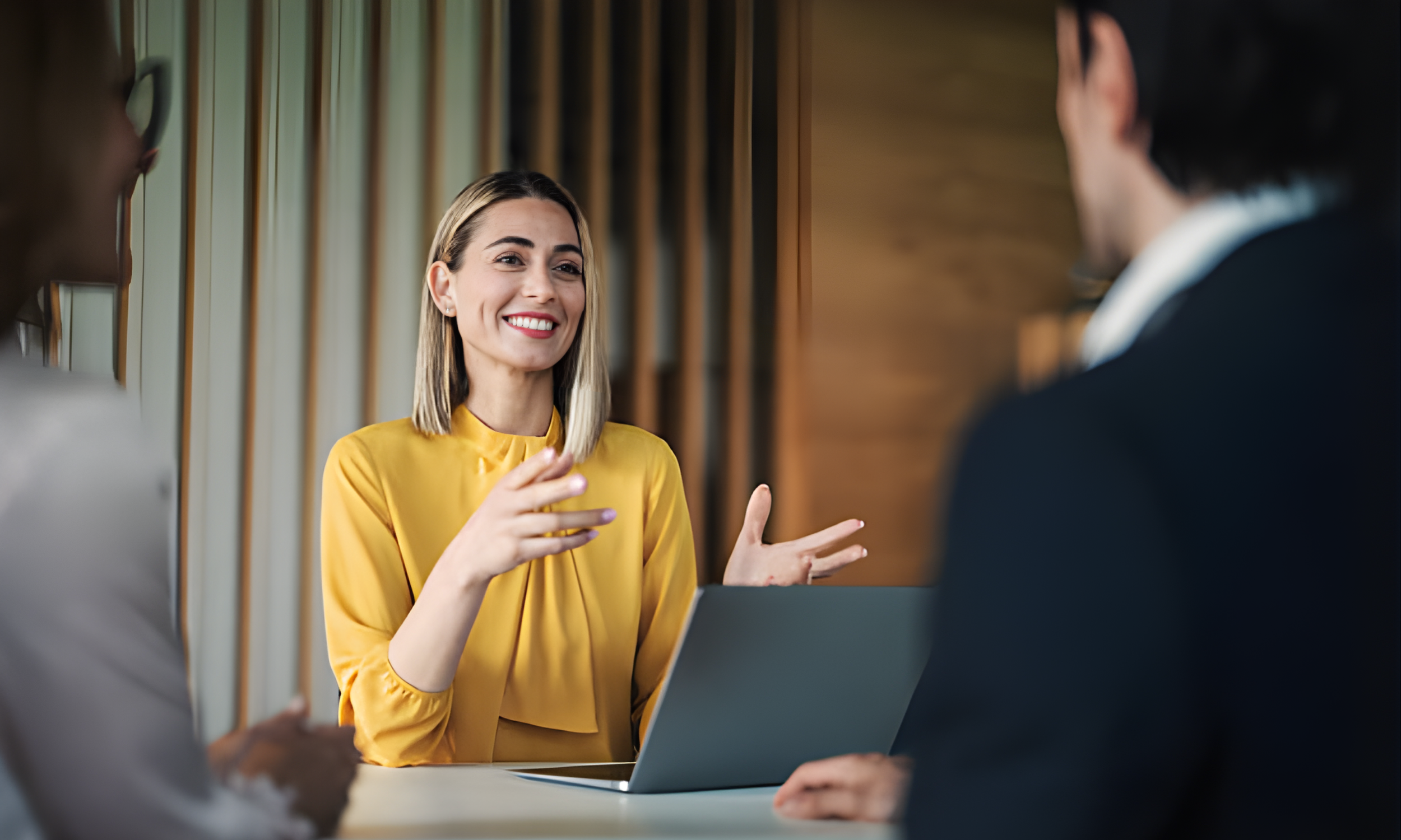 A woman in a yellow blouse smiling and speaking during a meeting with two other people, a man on the right and a woman on the left, in a modern office setting with wood paneling.
