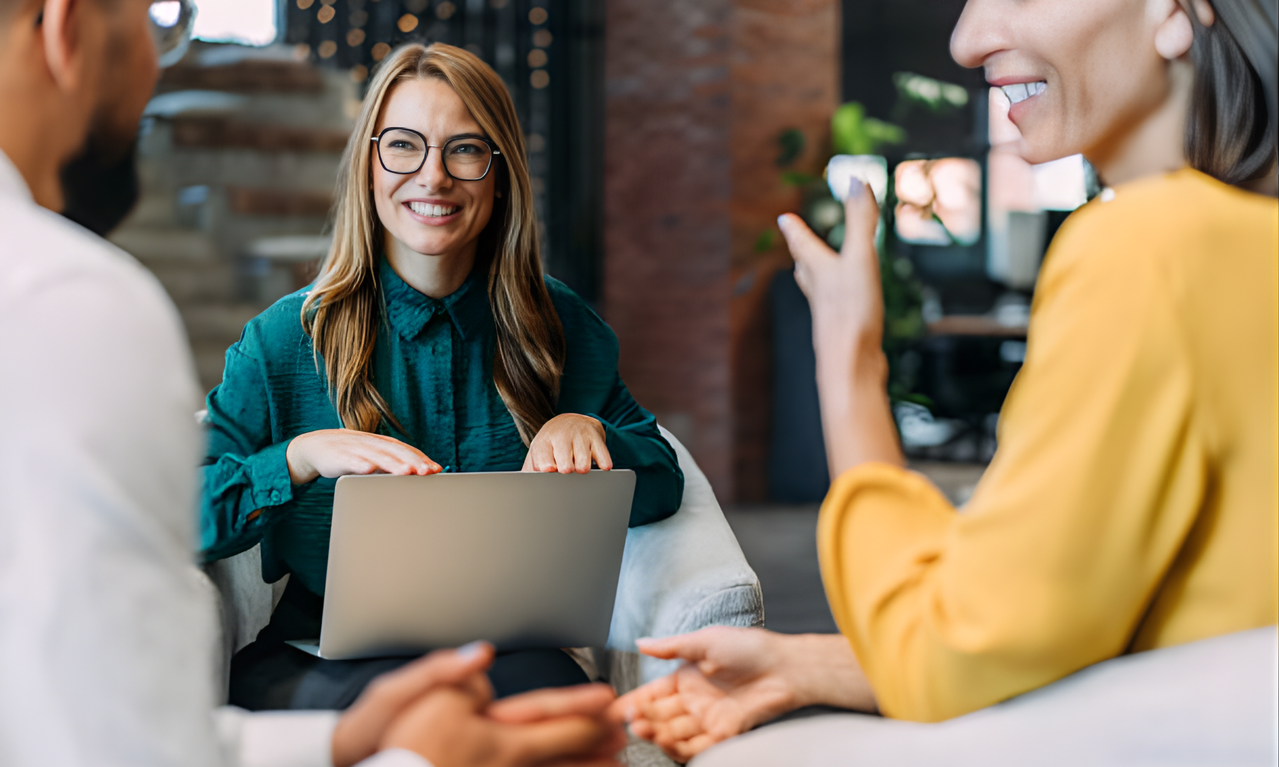 Three people in a professional setting engaged in a conversation, with a woman in glasses smiling and using a laptop, and two others partially visible, one gesturing with their hand and the other holding a phone.