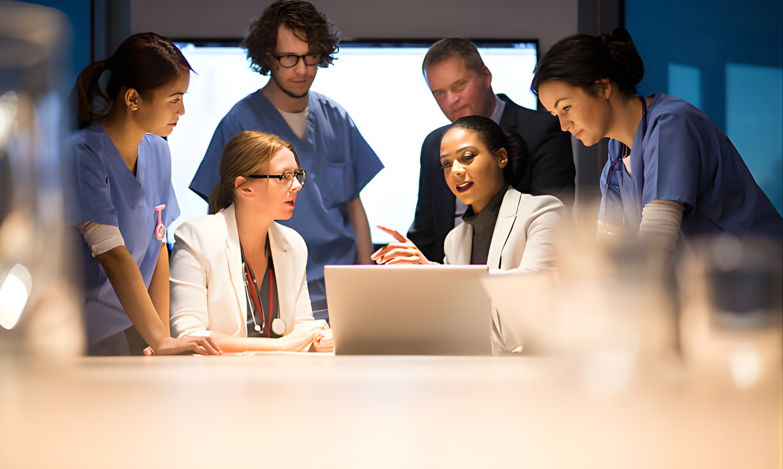 Group of medical professionals and a woman in a white blazer having a discussion around a laptop in a medical setting.