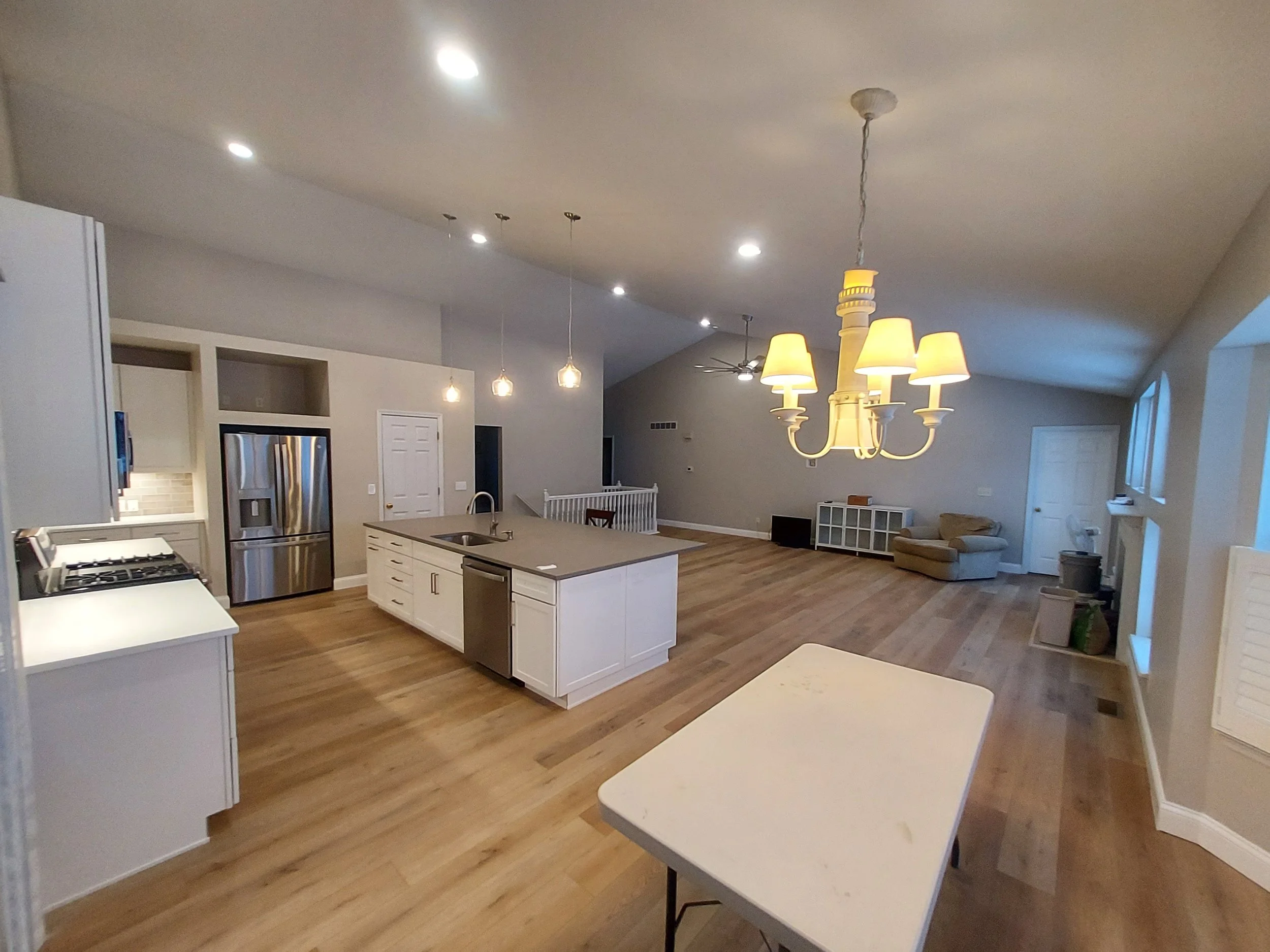 Open-concept kitchen and living room with hardwood floors, white cabinetry, stainless steel appliances, a large island with a sink, and a chandelier. The space includes a seating area with a beige armchair and a white storage unit, with windows letting in natural light.