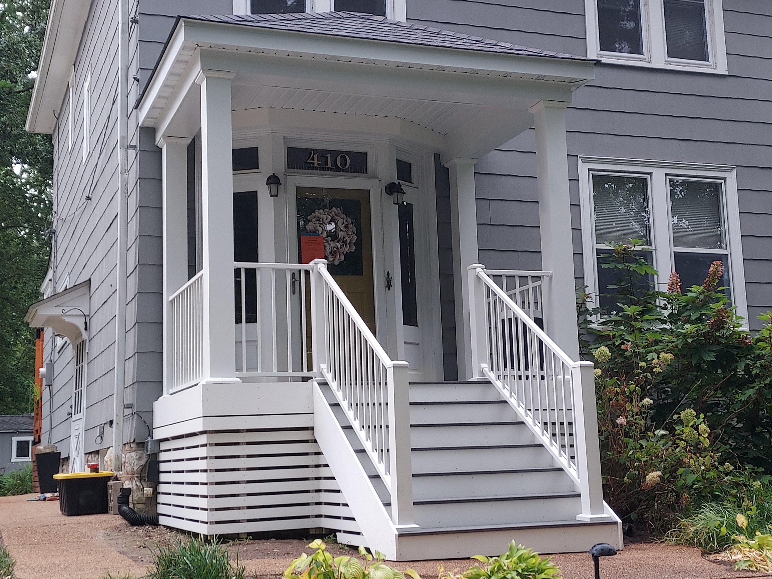 Front porch of a gray house with white railing and stairs, with lush green bushes and flowers on the right side.