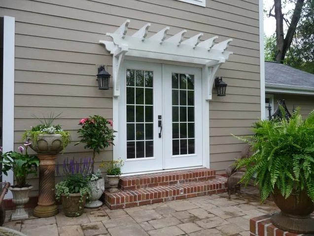 Exterior door with glass double doors, brick steps, and a white decorative trellis overhead. Flanked by black wall-mounted lanterns and surrounded by potted plants and greenery.