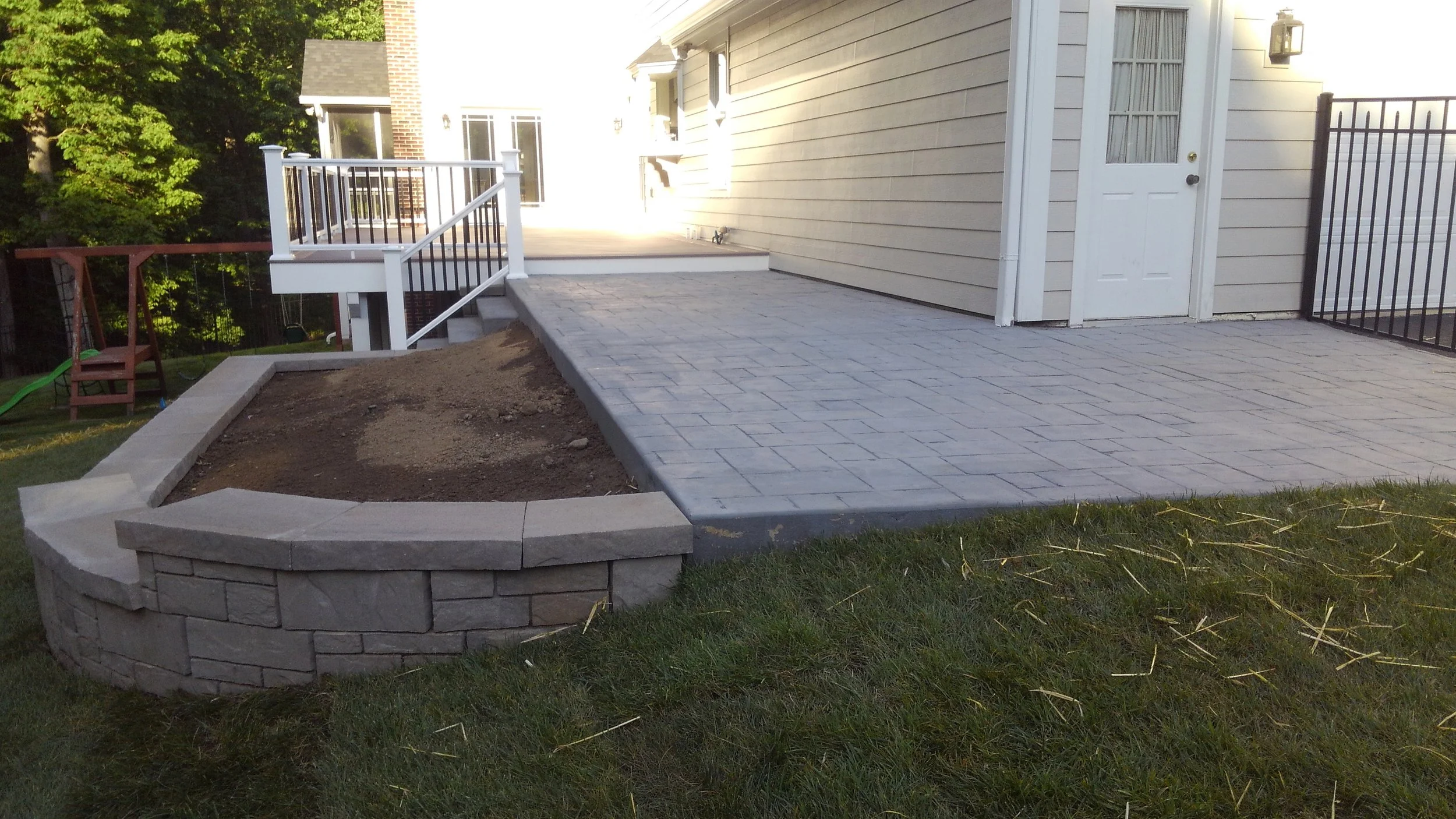 Backyard patio with newly paved and stamped concrete surface, a small curved stone retaining wall, and a white house with a door and windows, surrounded by green grass and trees.