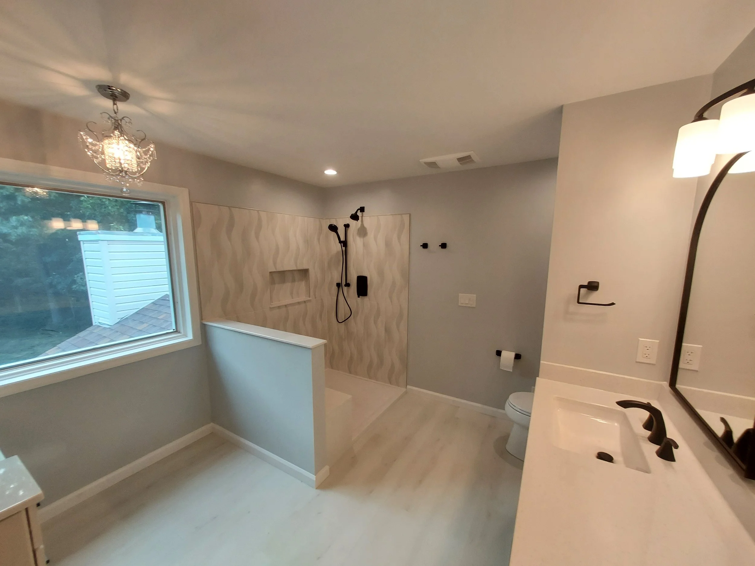 Modern bathroom with a large window, a chandelier, a walk-in shower with textured wall tiles, black fixtures, a toilet, and a white vanity with a black faucet.