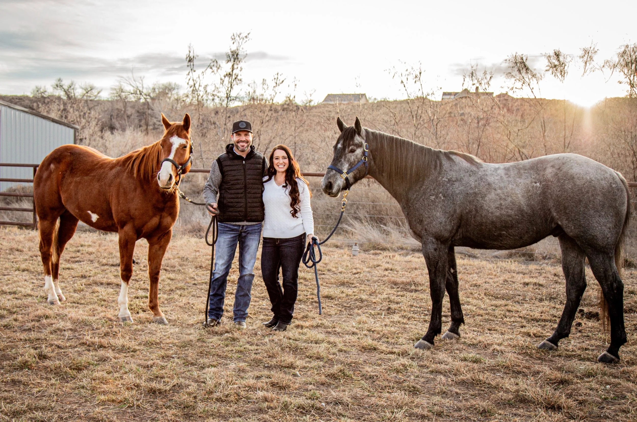 Happy couple standing with two horses outdoors during sunset.