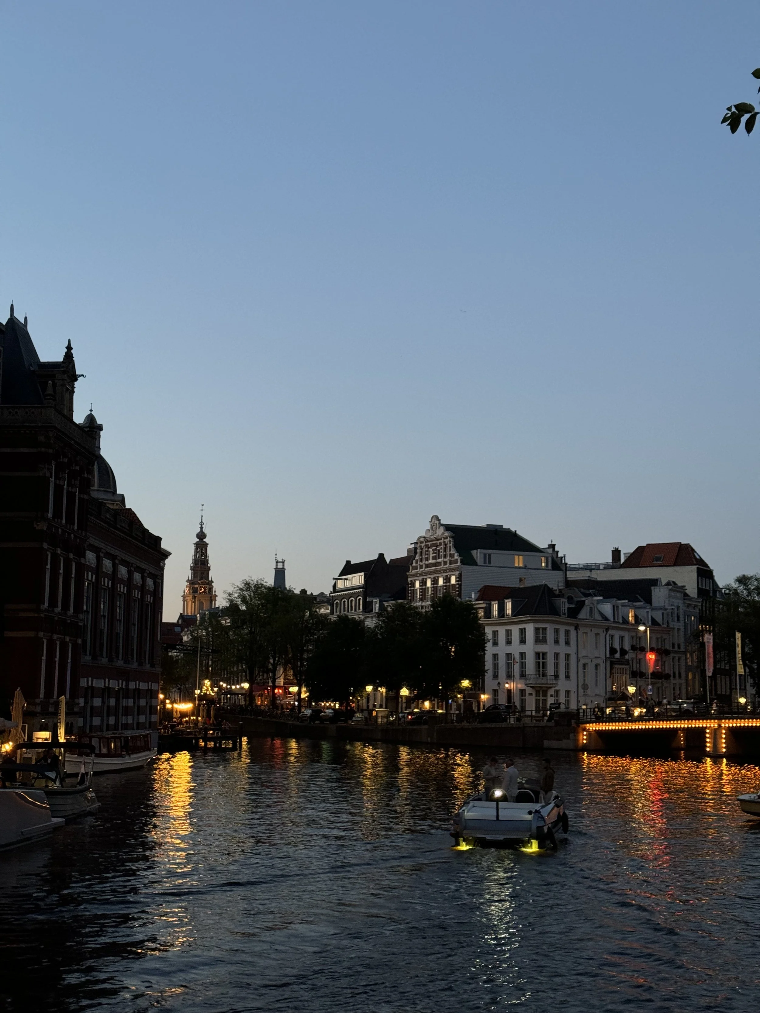 Cityscape at dusk with a canal, boats, historic buildings illuminated, and a clear sky.