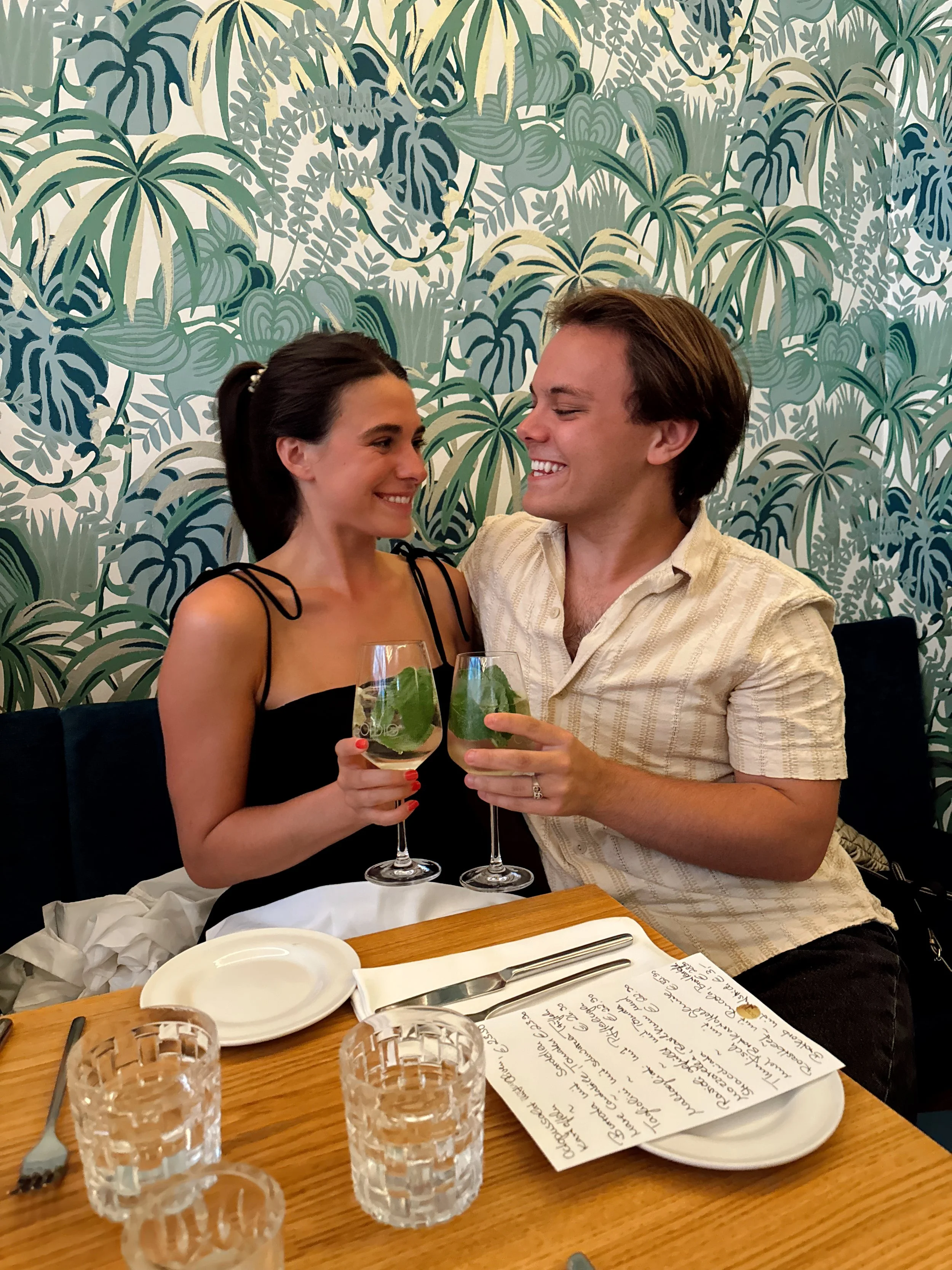 A young couple sharing a toast with cocktails at a restaurant table, smiling at each other. The table has glasses, plates, and a handwritten menu. The background features leafy patterned wallpaper.
