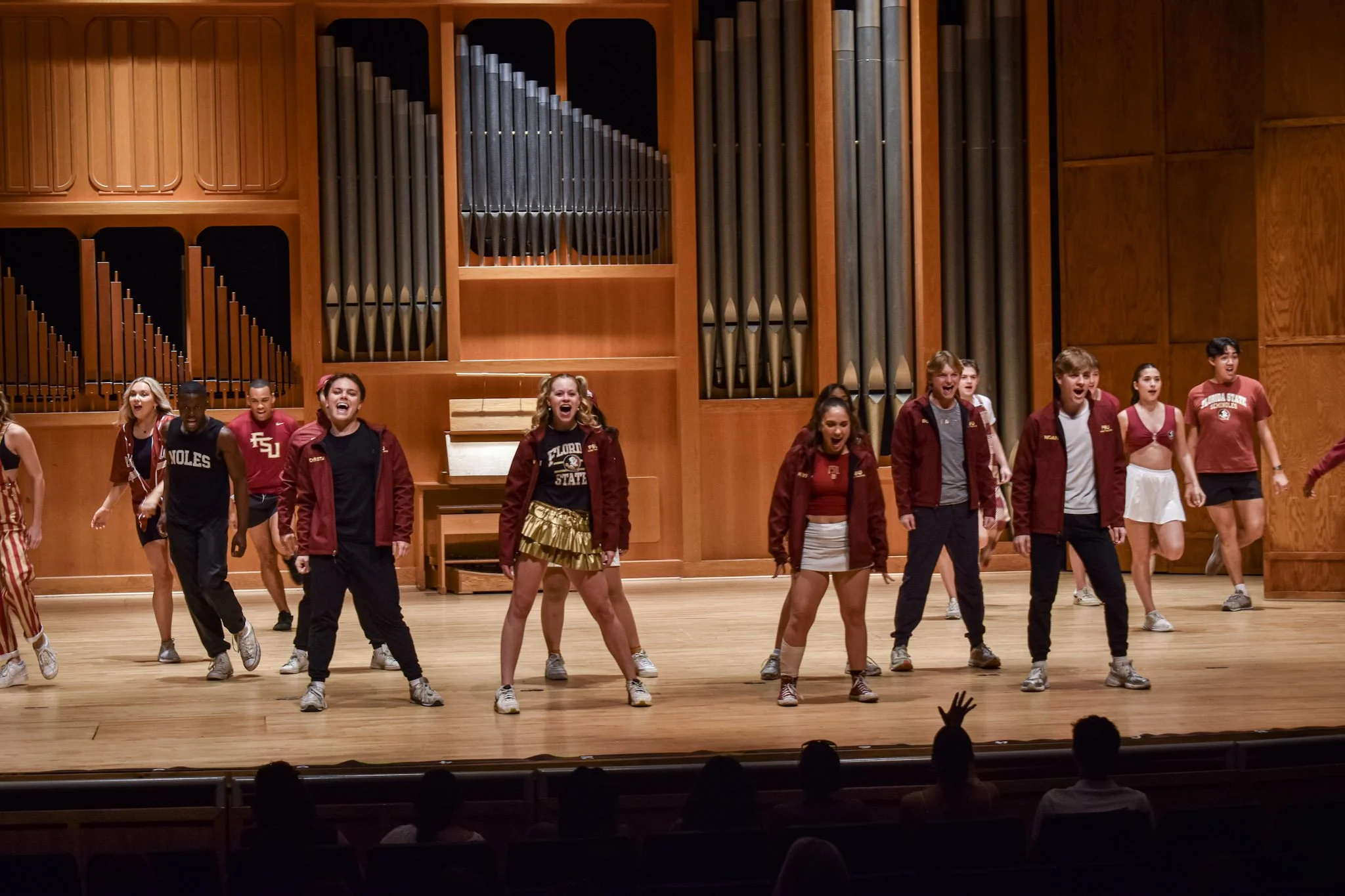 Group of teenagers performing a dance or cheer routine on stage in a concert hall, with a large pipe organ in the background.
