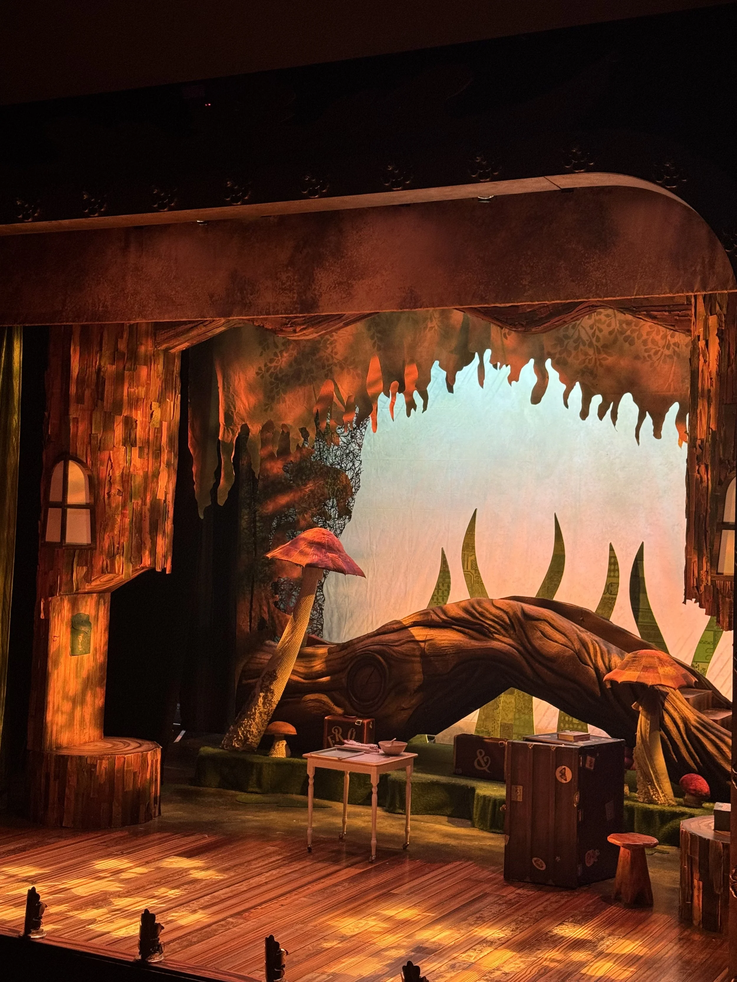 The stage is decorated to resemble a whimsical forest scene, with large tree-like structures, oversized mushrooms, and a backdrop depicting trees and nature. There are some props like a small table and stools placed on the stage.