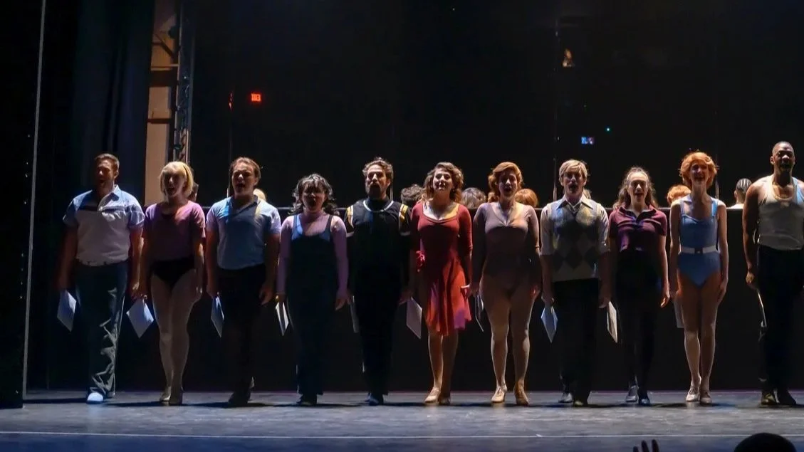 Stage with 16 performers standing in a line, lit by blue and yellow stage lights, with a dark background.