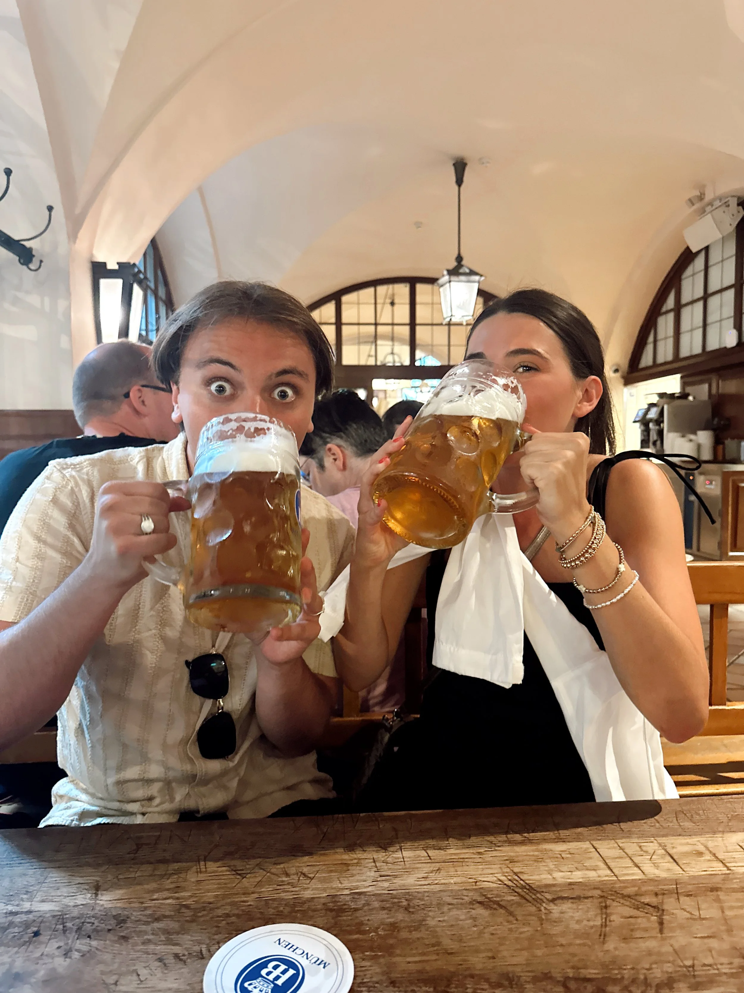 Two friends enjoying beers at a restaurant, smiling and holding large mugs of beer to their mouths.