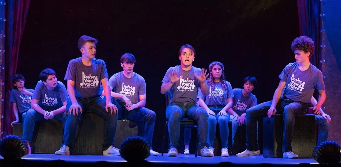Children on stage wearing matching t-shirts, with one child speaking and others sitting and listening during a performance.