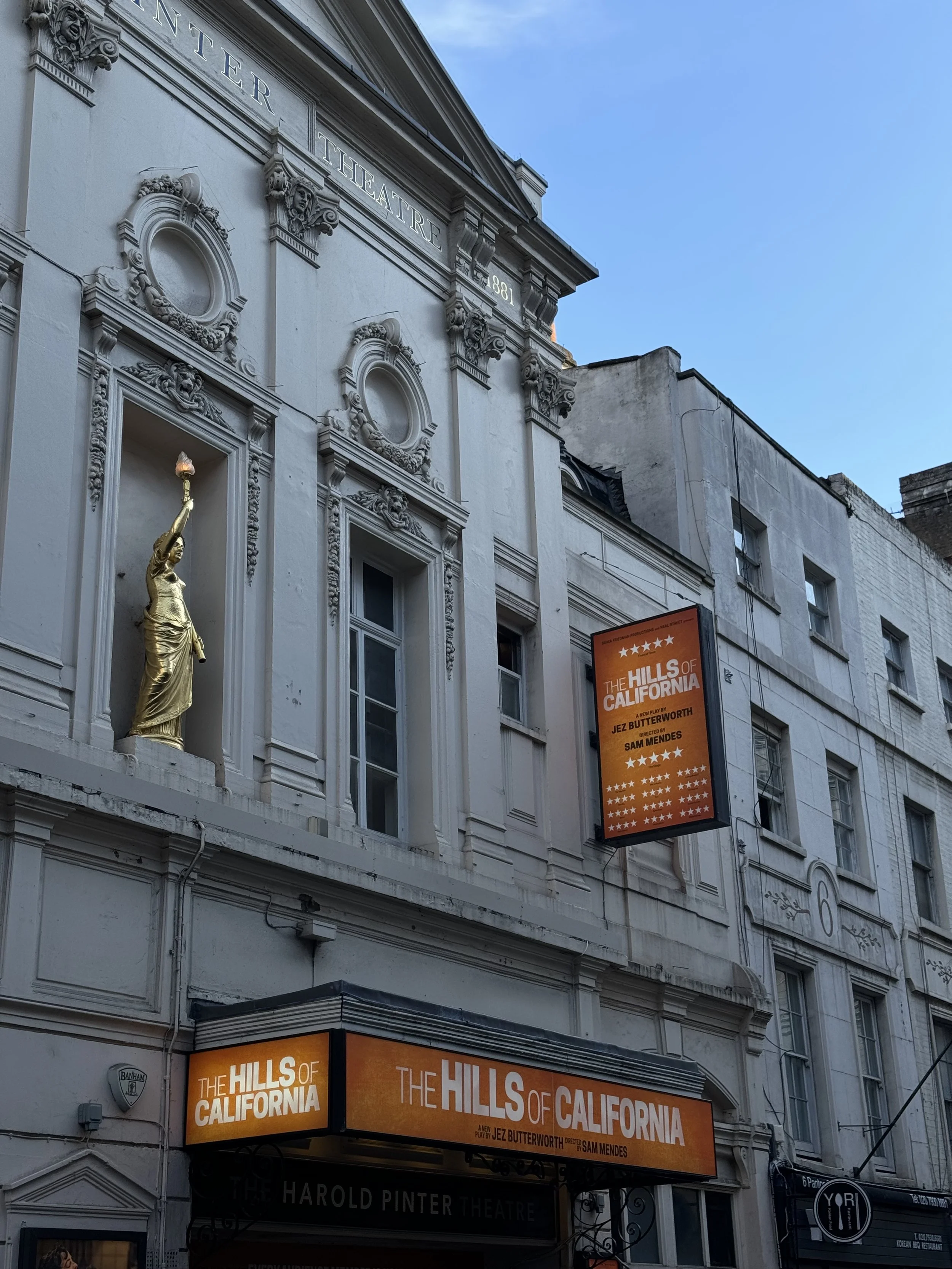 Exterior of the Harold Pinter Theatre with orange signage for the play "The Hills of California" by Jez Butterworth, directed by Sam Mendes. A golden statue of a woman holding a torch stands on a ledge above the entrance. The building has classical a
