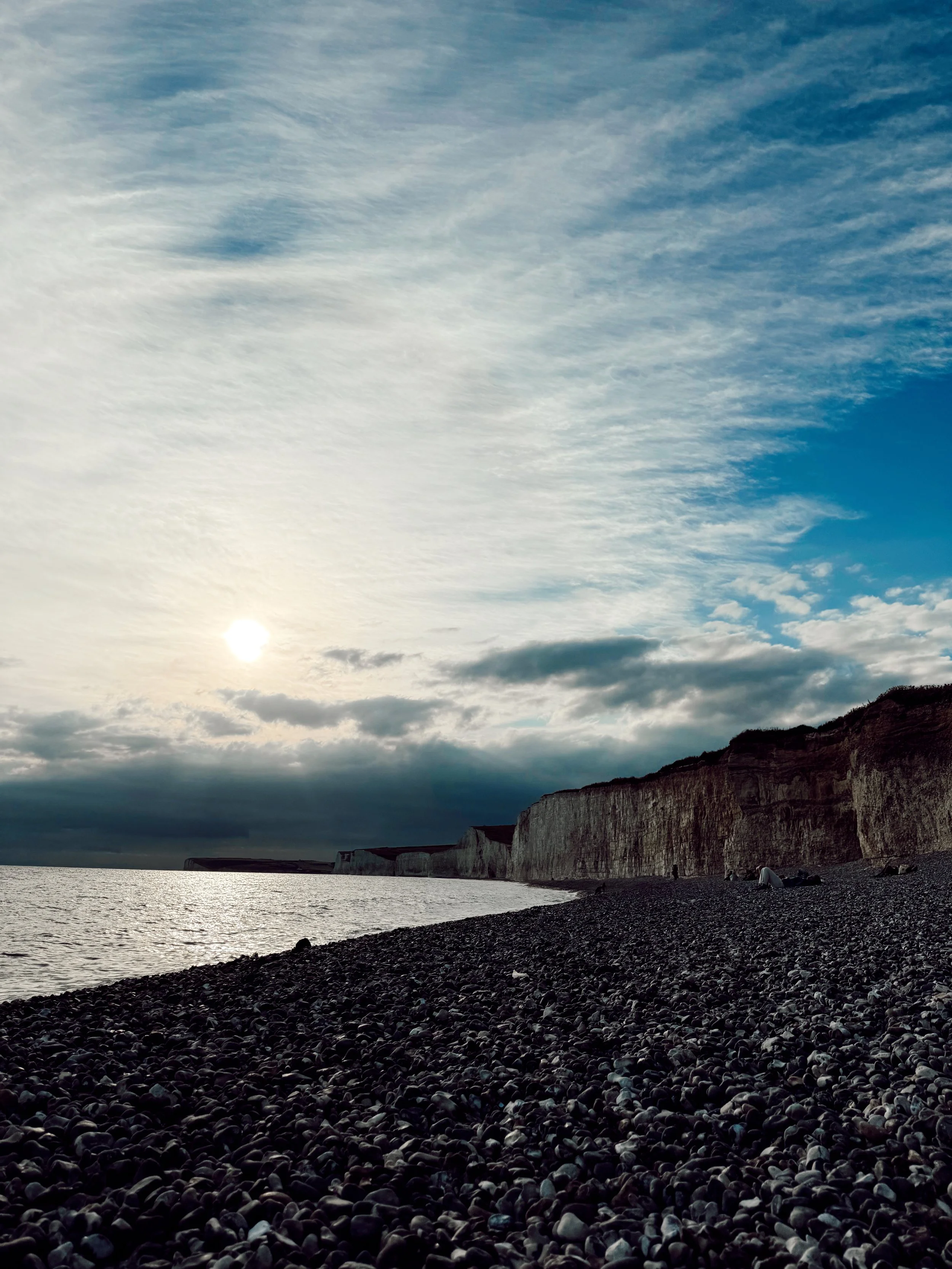 A rocky beach with white chalk cliffs in the distance and a cloudy sky with the sun partially obscured by clouds.