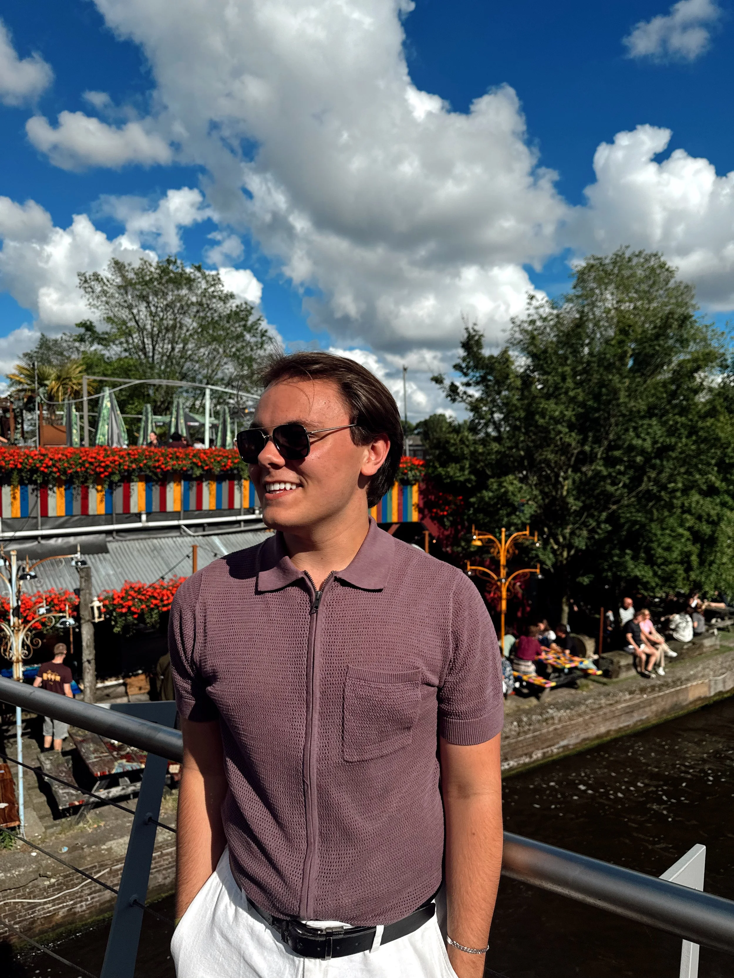 A young man with dark hair, wearing a maroon short-sleeved shirt, white pants, and sunglasses, standing outdoors near a river with a lively background of trees, a colorful building, and people relaxing on benches during a sunny day with blue skies an