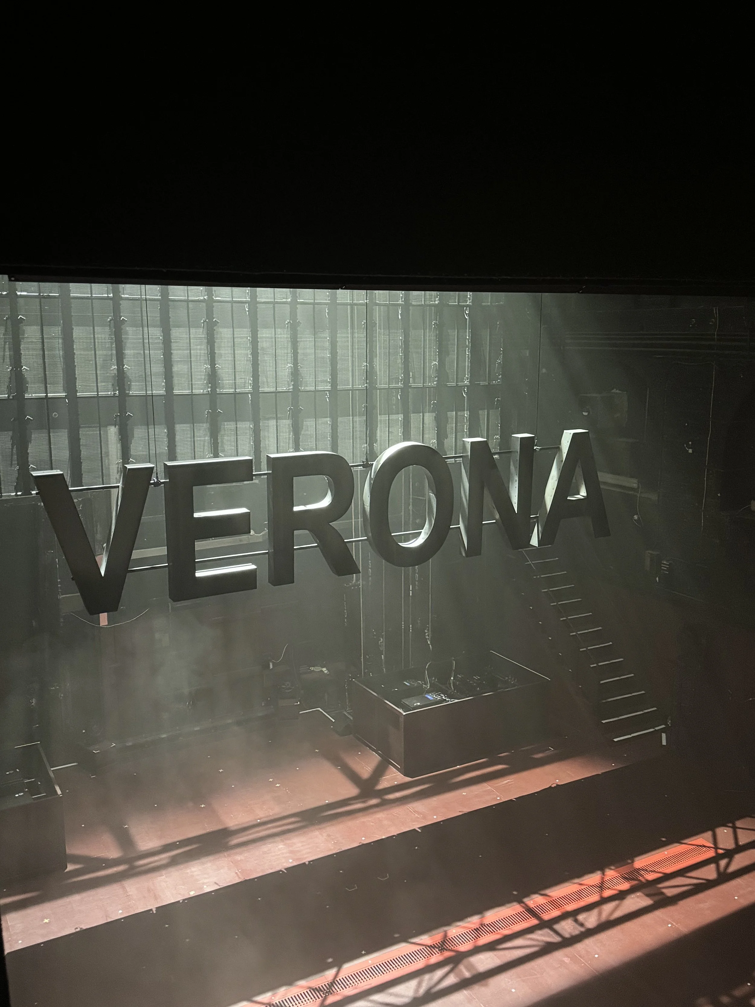 Inside view of a stage with large 'VERONA' sign, stairs on the right, and sunlight streaming through large glass windows.
