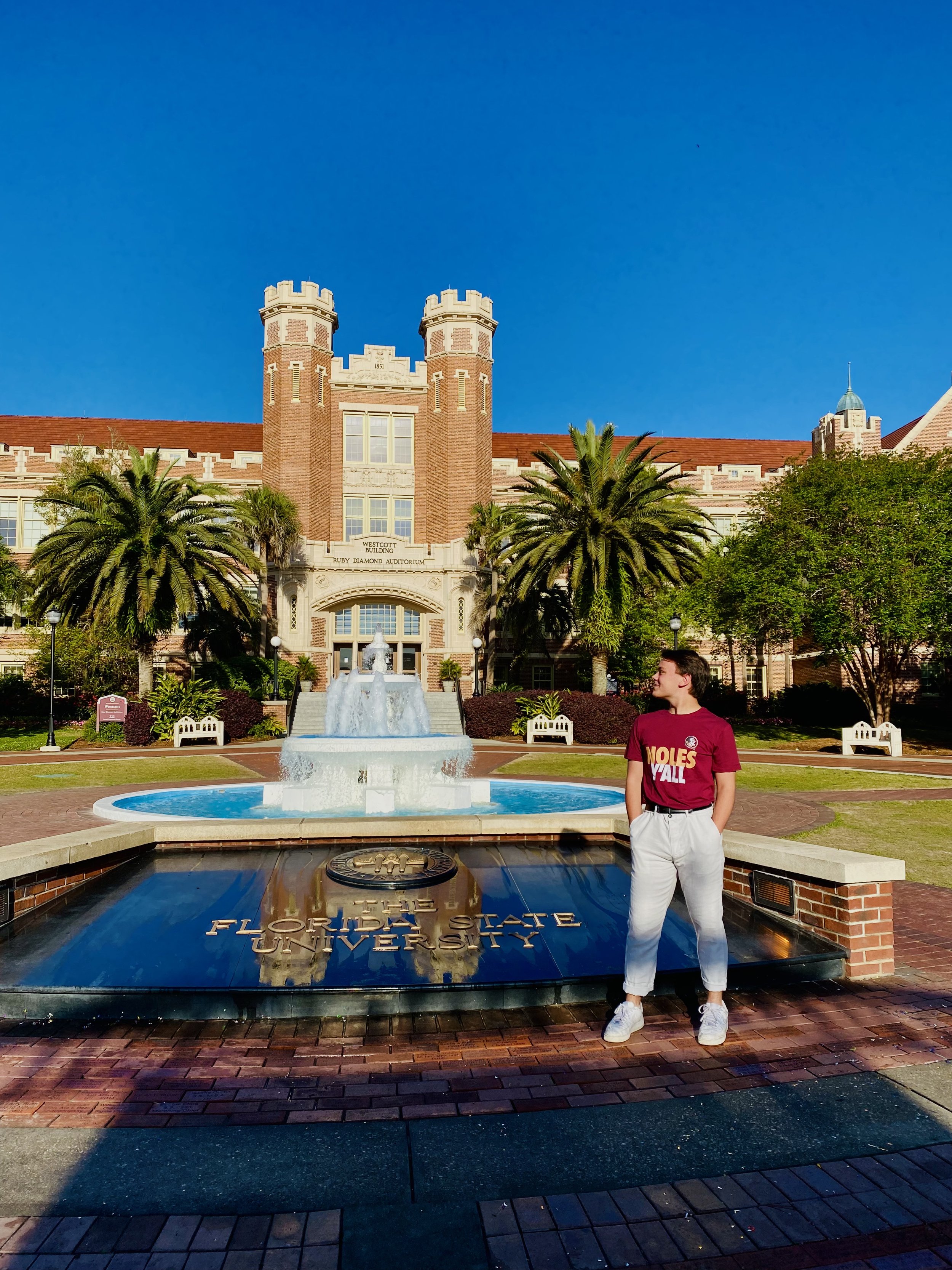 A person standing outside Florida State University's Ruby Diamond Auditorium, with palm trees, a fountain, and a brick building in the background on a clear, sunny day.
