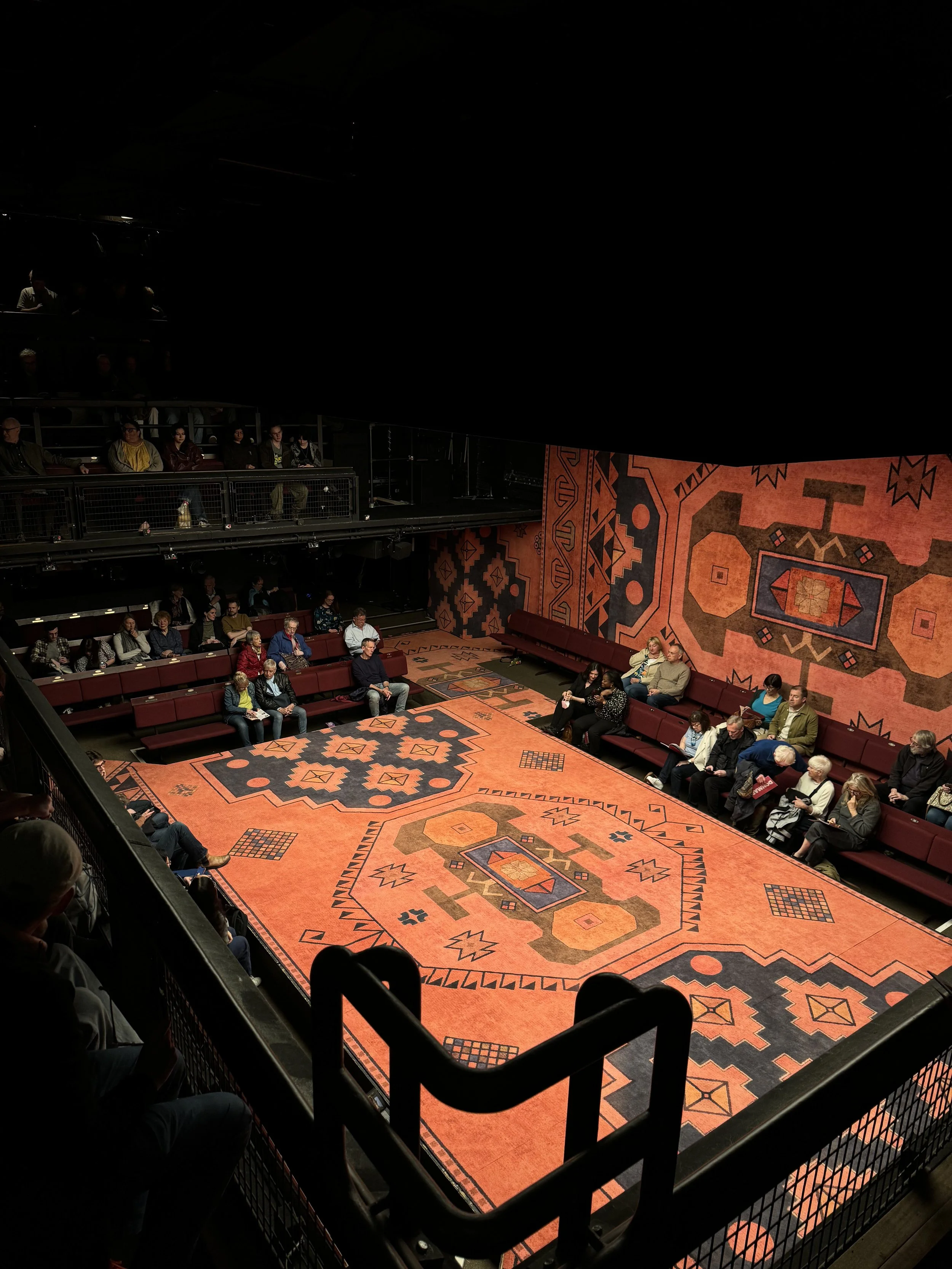 Audience seated in a theater with a decorated orange and black patterned floor and walls, watching a performance.