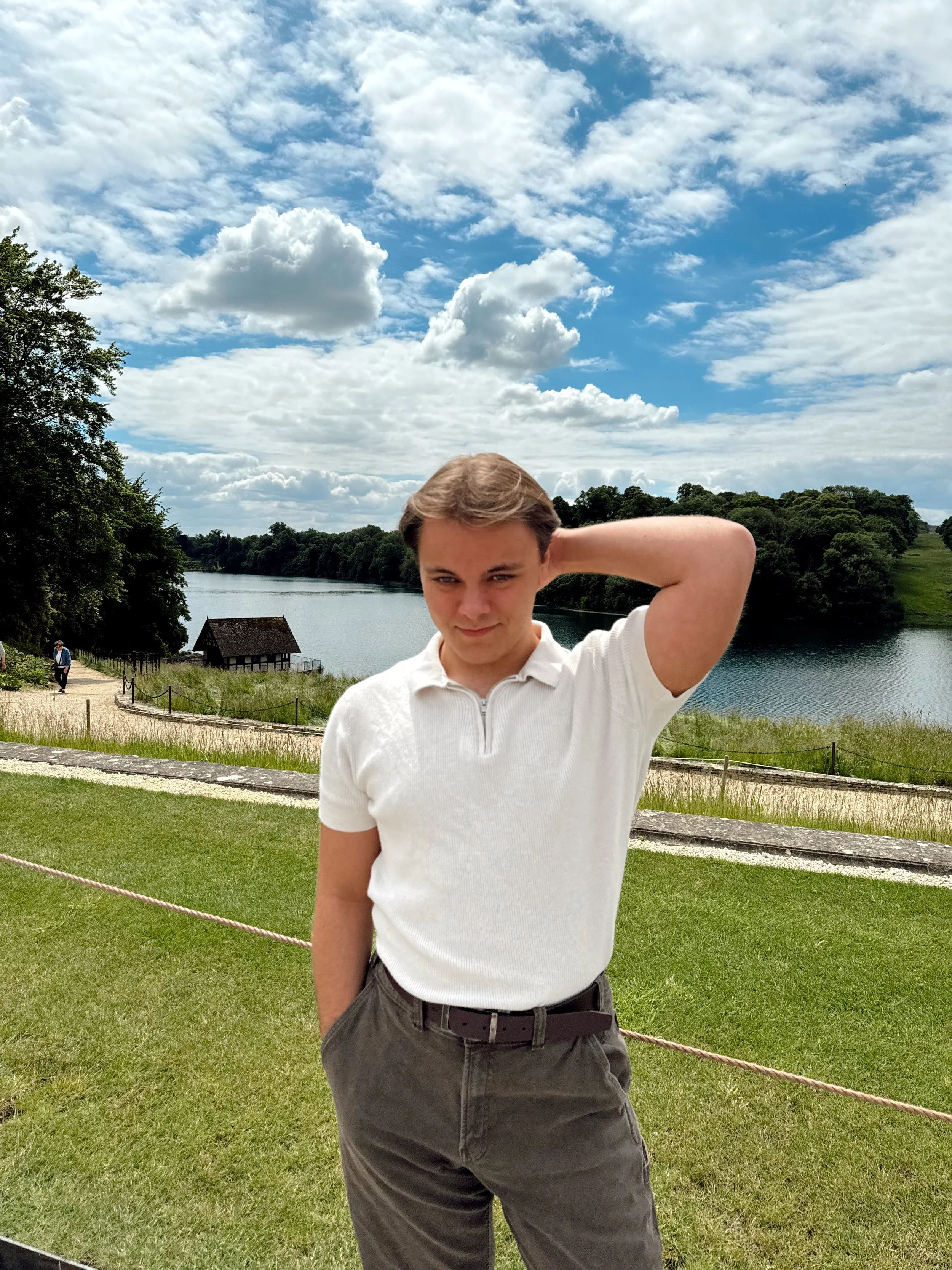 A young man in a white polo shirt and dark gray pants standing outdoors near a lake, with trees, a small building, and a partly cloudy sky in the background.