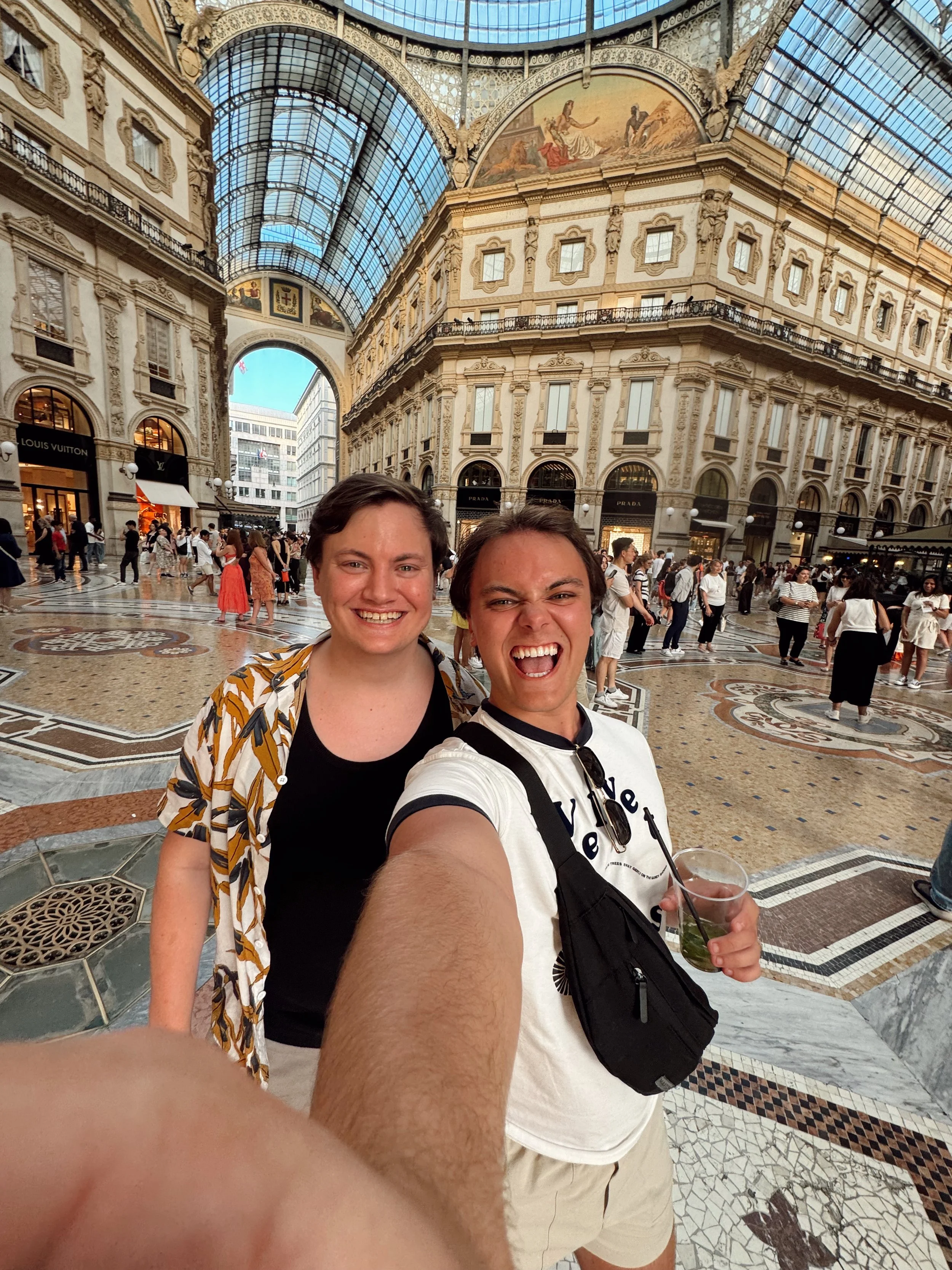 Two brothers taking a selfie inside a grand shopping arcade with ornate architecture and a glass ceiling. 