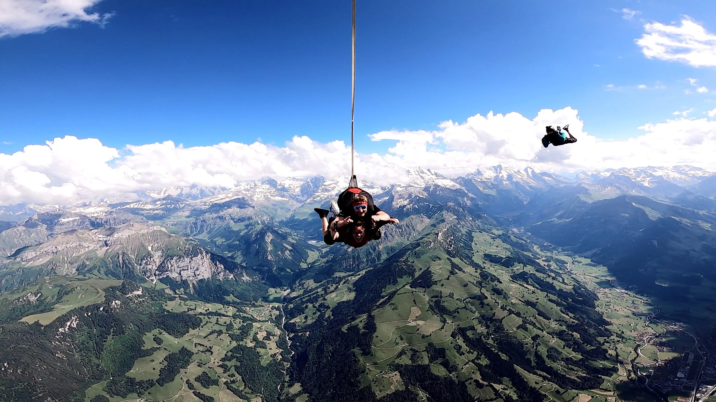 Two people hang from a harness high above green mountains and valleys, with snow-capped peaks in the distance, under a partly cloudy blue sky.