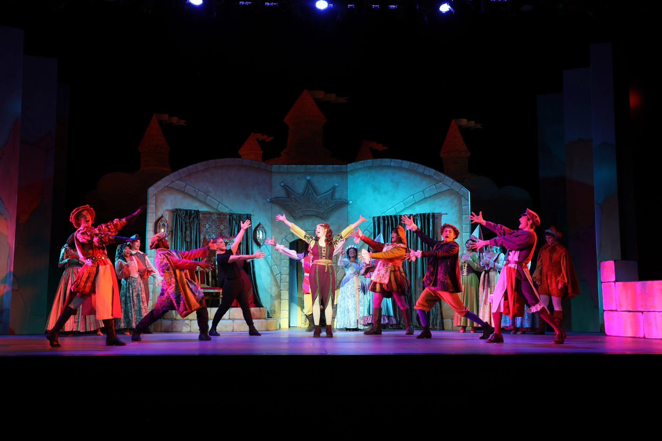 Theatrical stage performance of a cast, dressed in colorful costumes, with some in the foreground extending their arms, set against a castle backdrop with turrets and banners.