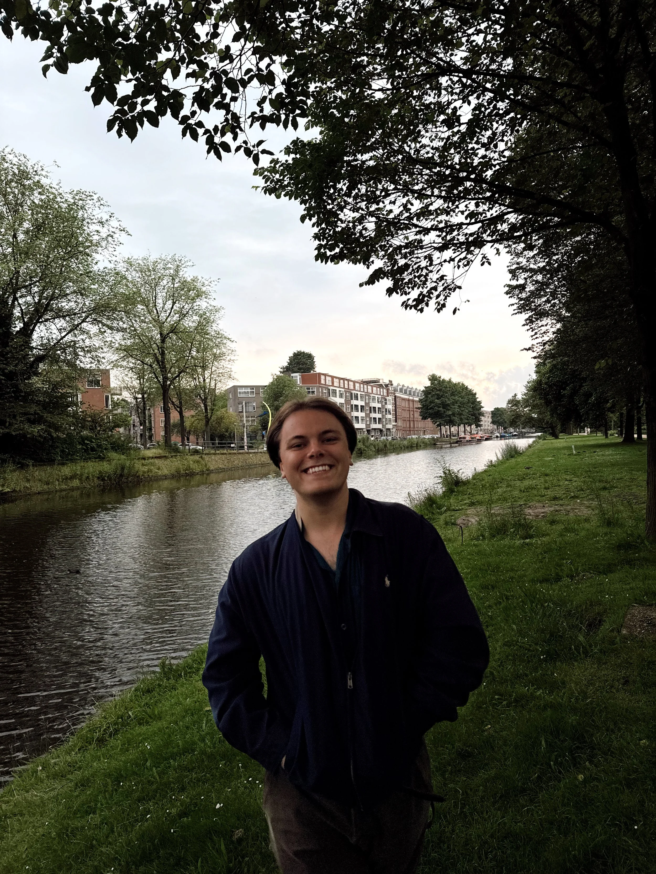 A young man with short dark hair smiling while standing outdoors by a canal, surrounded by trees and modern buildings in the background, on a cloudy day.