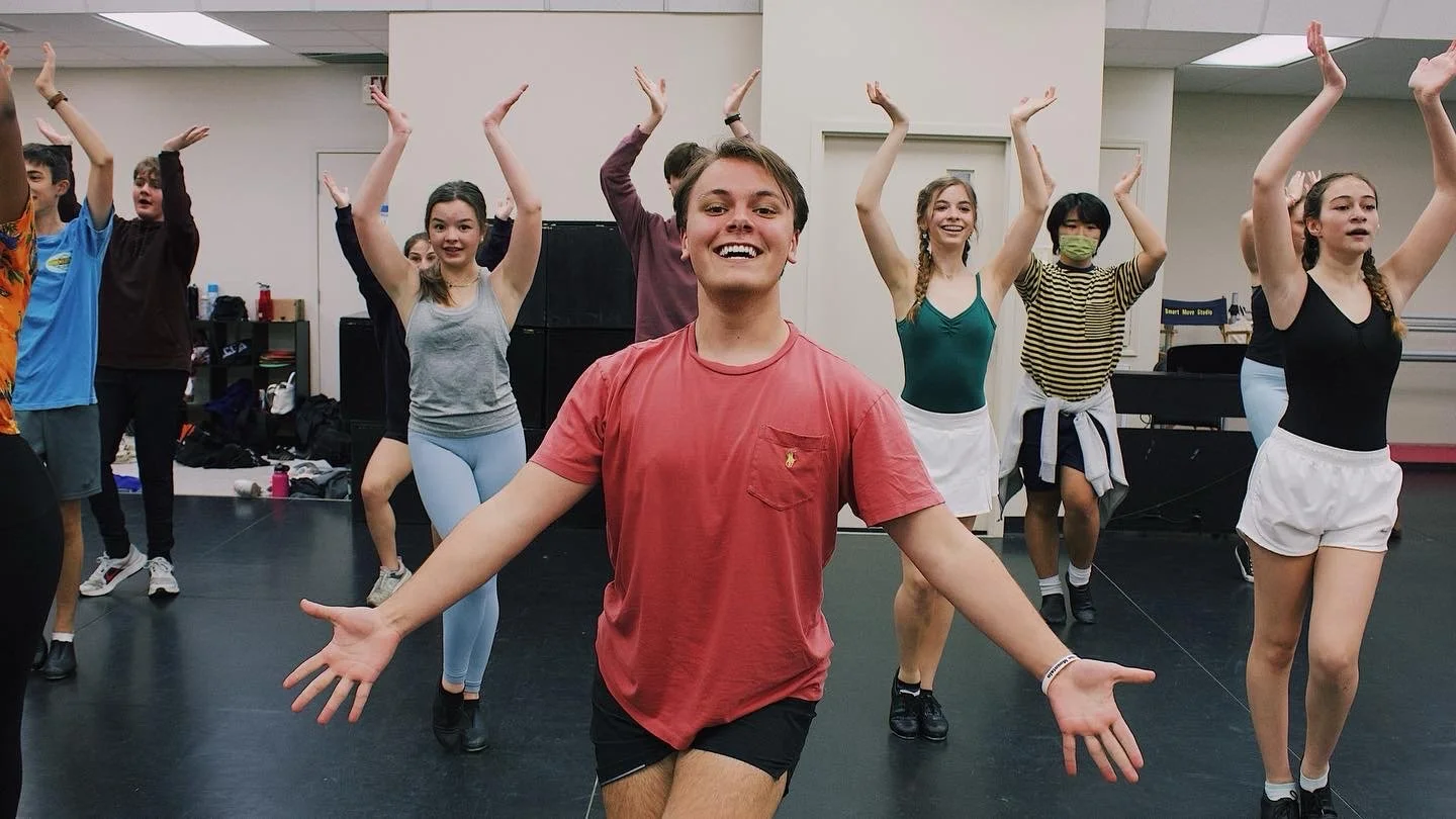Group of young dancers practicing in a studio, some raising their arms, with a smiling person in front facing the camera.