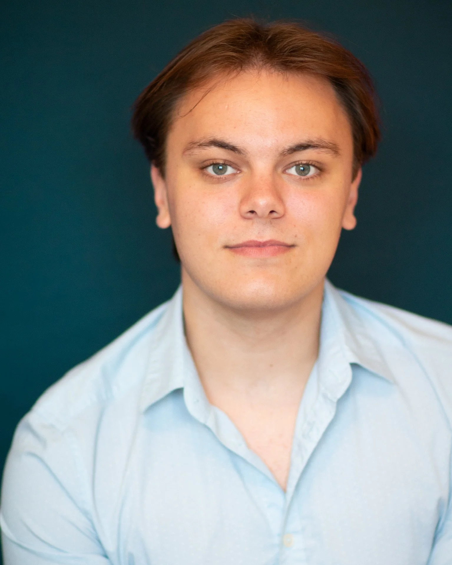 Headshot of a young man with light brown hair wearing a light blue collared shirt, facing forward against a dark teal background.