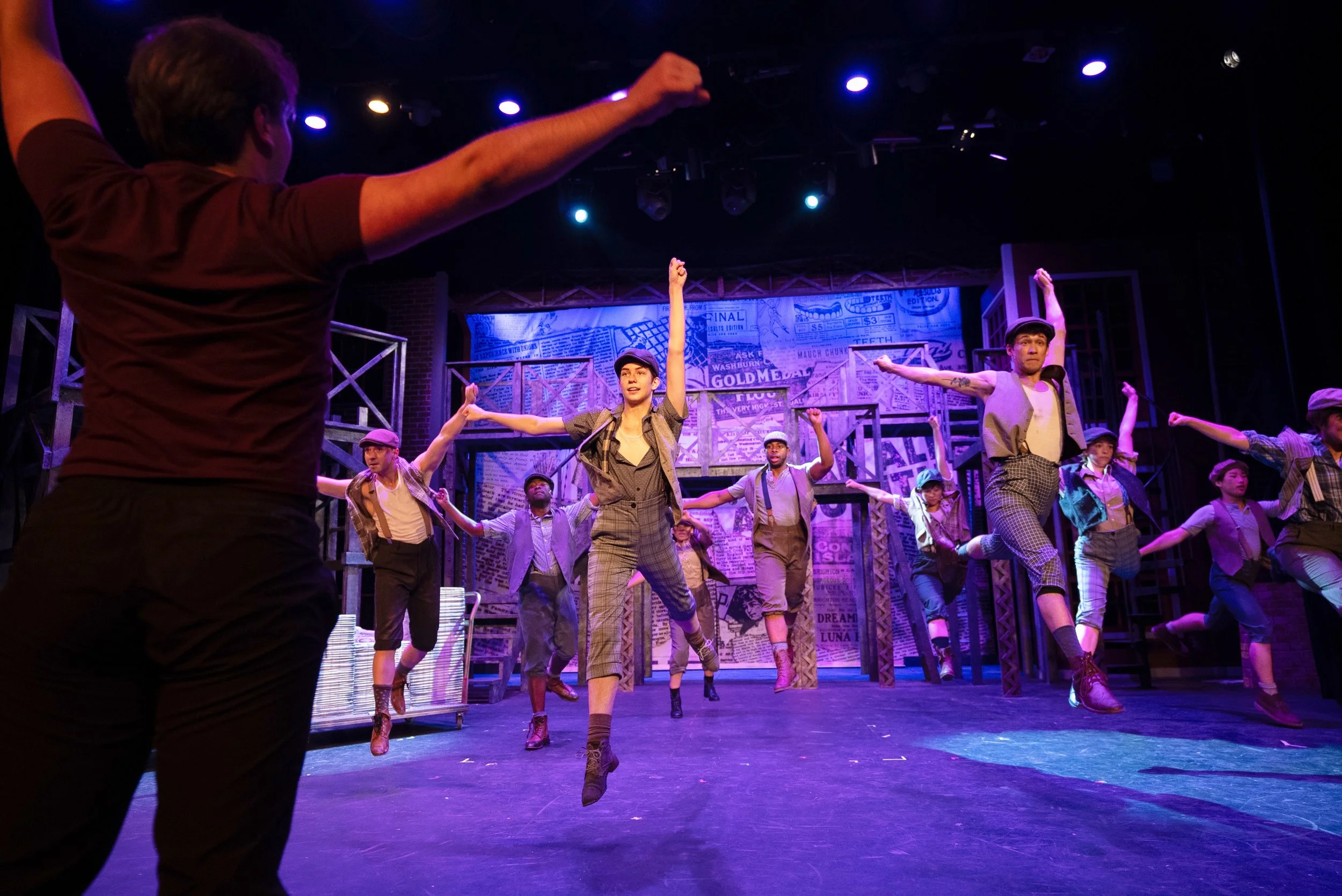 Theater stage with performers dancing in choreographed positions, wearing vintage attire, lit with blue lights, with a backdrop of newspaper-themed set pieces.