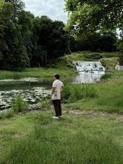 Person standing near a pond with a small waterfall, surrounded by green trees and grass.