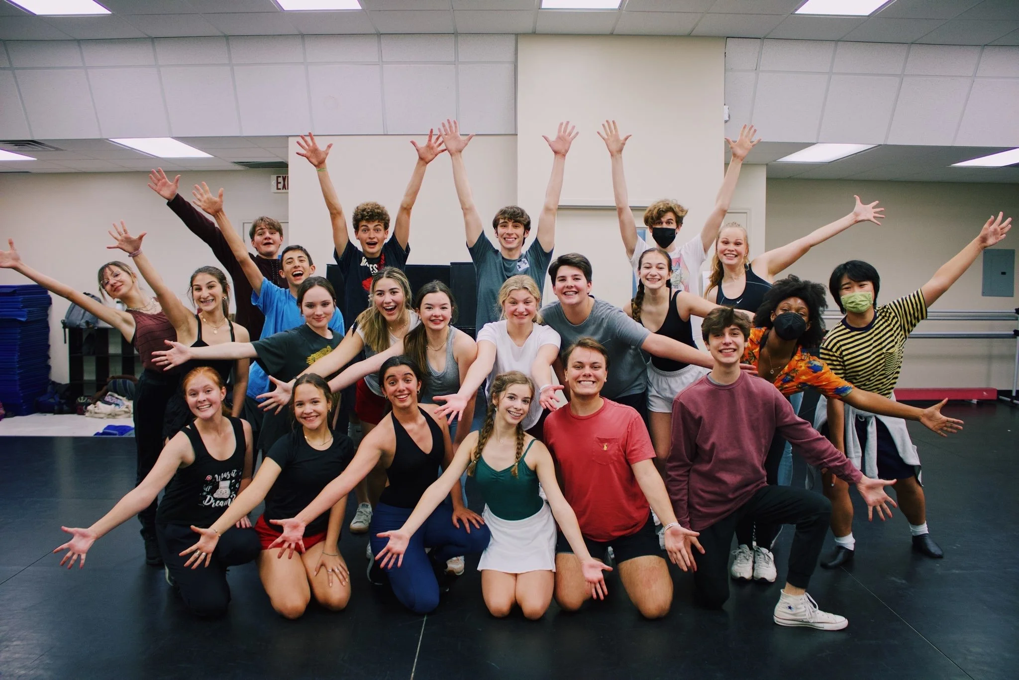 A group of teenagers, some wearing masks, are posing with their arms outstretched in a dance studio or rehearsal space with a black floor, white walls, and a drop ceiling. They are smiling and appear happy.