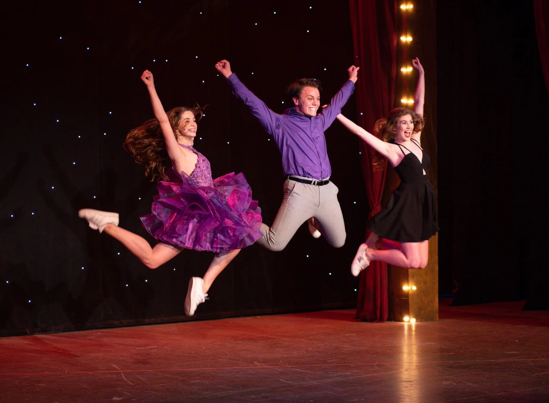 Three young performers jumping in mid-air on stage with a dark background and red curtains.